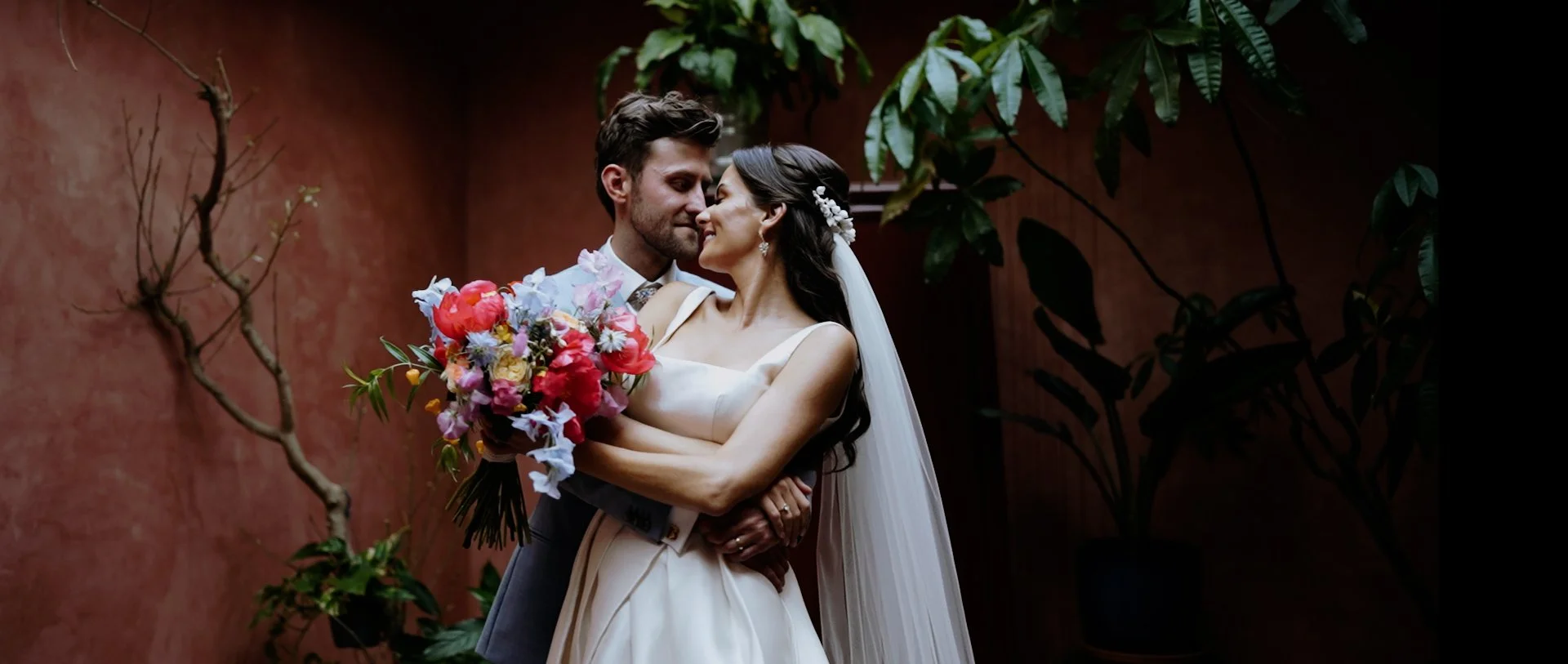 Bride and groom sharing a romantic moment indoors, with the bride holding a colorful bouquet of flowers, surrounded by green plants and a textured red wall.