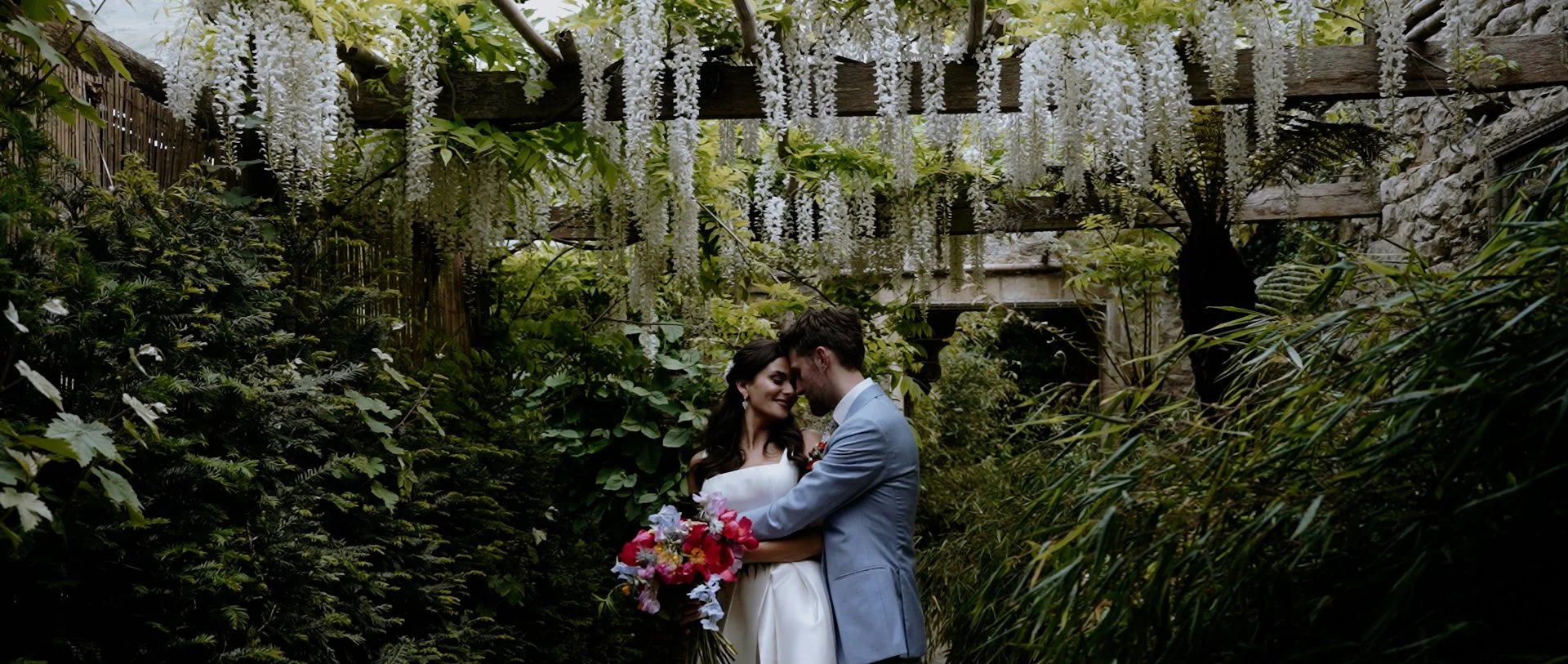 A bride and groom embrace in a lush, green garden with hanging white flowers overhead.