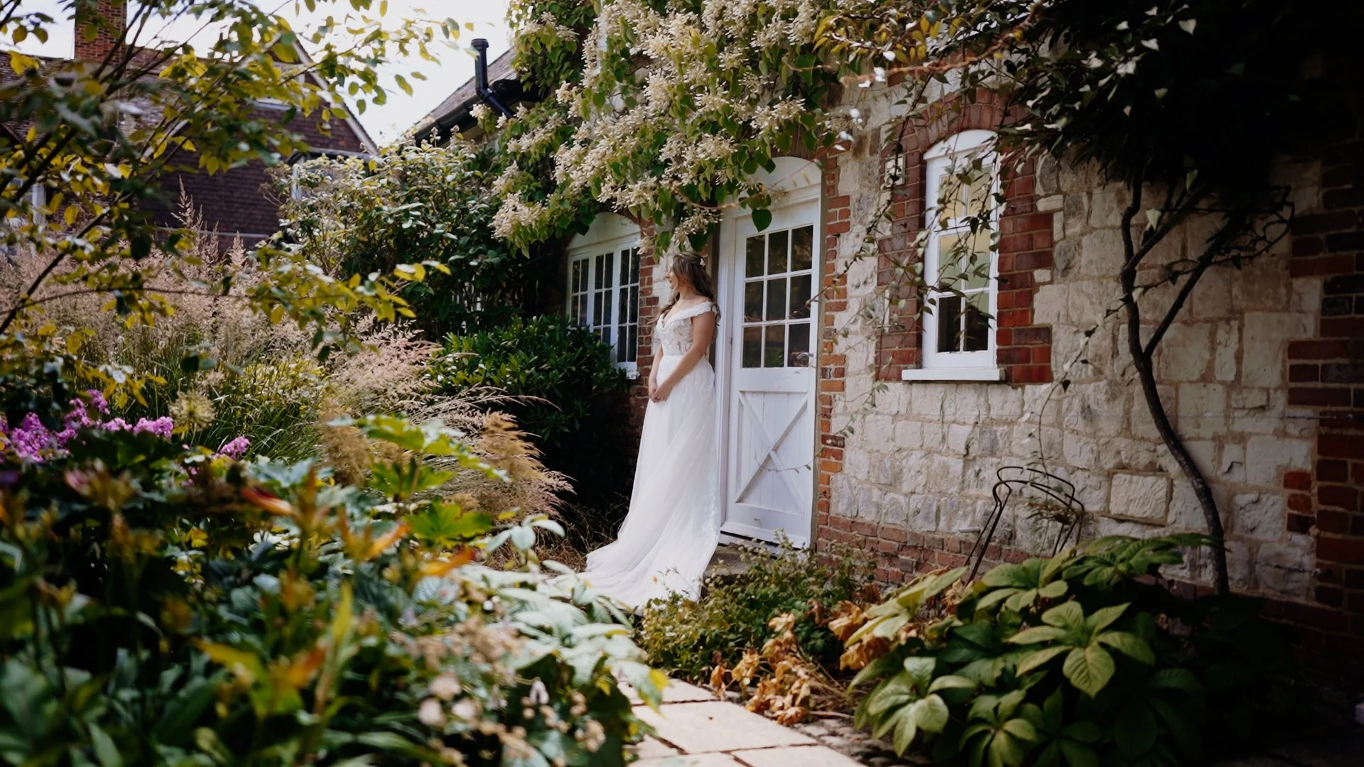 Woman in white dress standing outside a stone house surrounded by greenery and flowers.