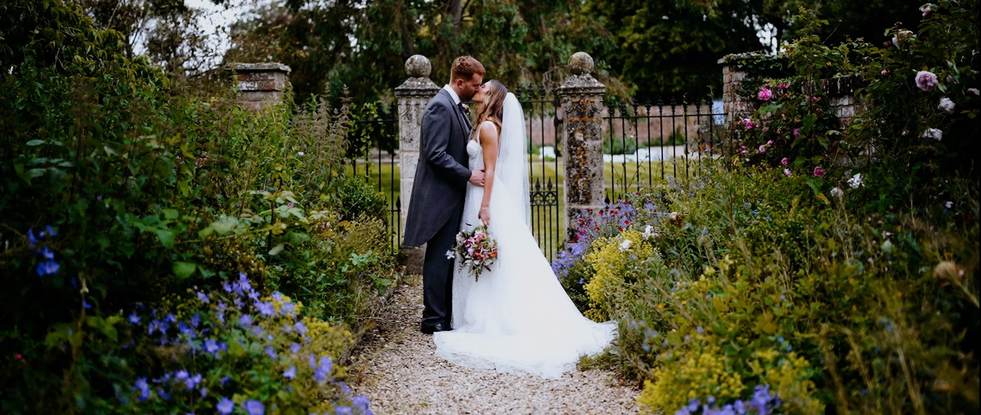 A bride and groom kiss in a garden surrounded by colorful flowers, with a stone fence and lush trees in the background.