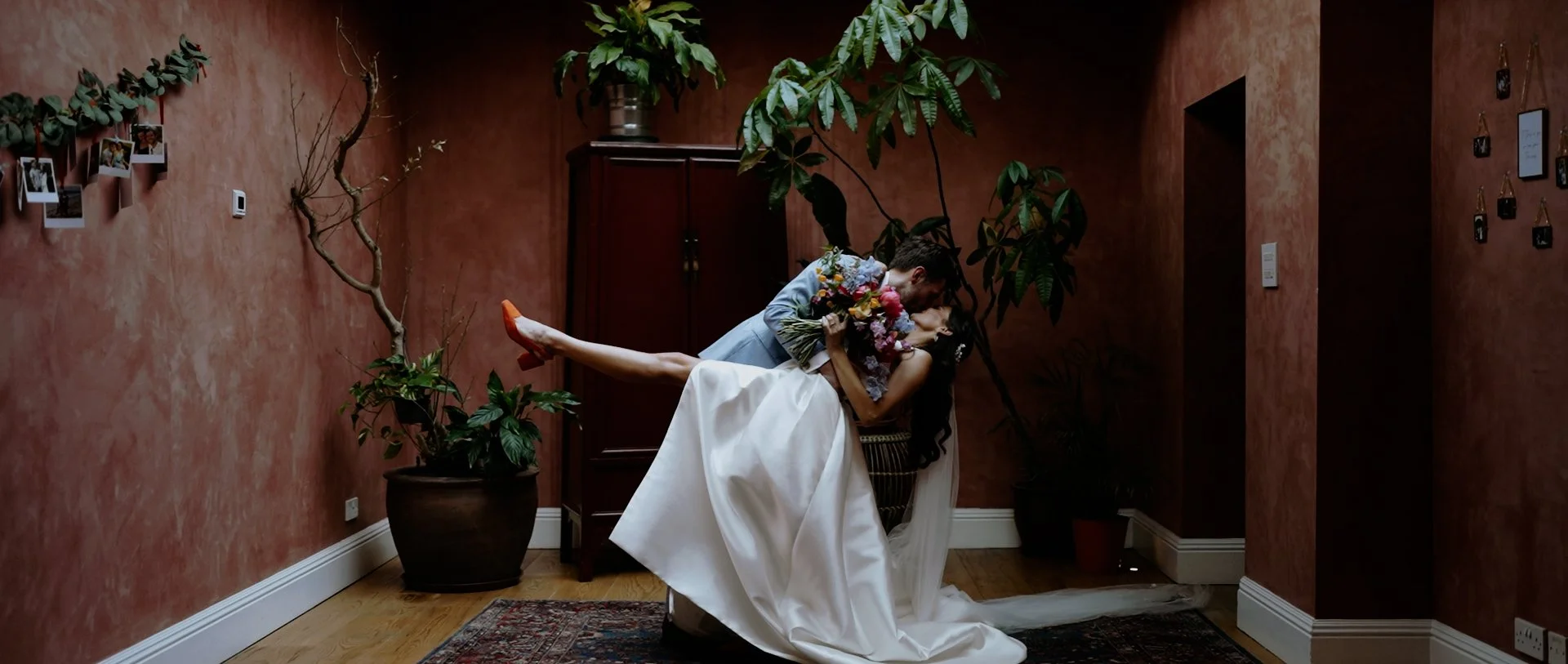 A bride and groom in a warm, indoor setting with red textured walls and large potted plants, where the groom is leaning over and holding the bride, who is holding a bouquet of flowers, as they share a joyful moment.