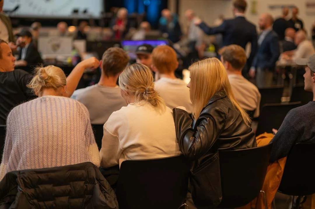 Folk deltar i en konferanse eller seminar, satt ved bord, mens andre snakker og går rundt i bakgrunnen.