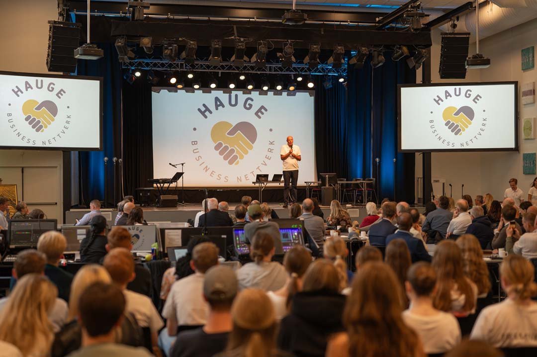En konferansesal med mange deltakere som ser på en taler på scenen, med to store skjermer som viser logoen til Hauge Business Nettverk, som er to hender som danner en hjerteform.