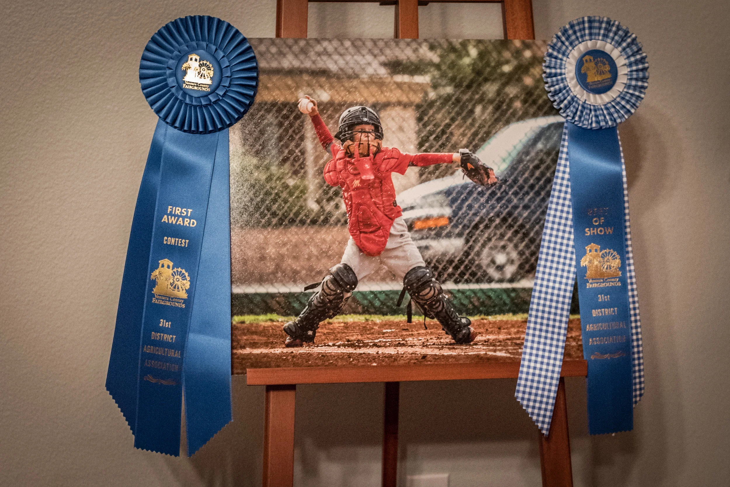 A framed photograph of a young baseball catcher, wearing a red jersey and catching gear, making a diving catch on a dirt field with a chain-link fence and a parked car in the background. Two blue ribbon awards are hanging on the sides of the photograph.