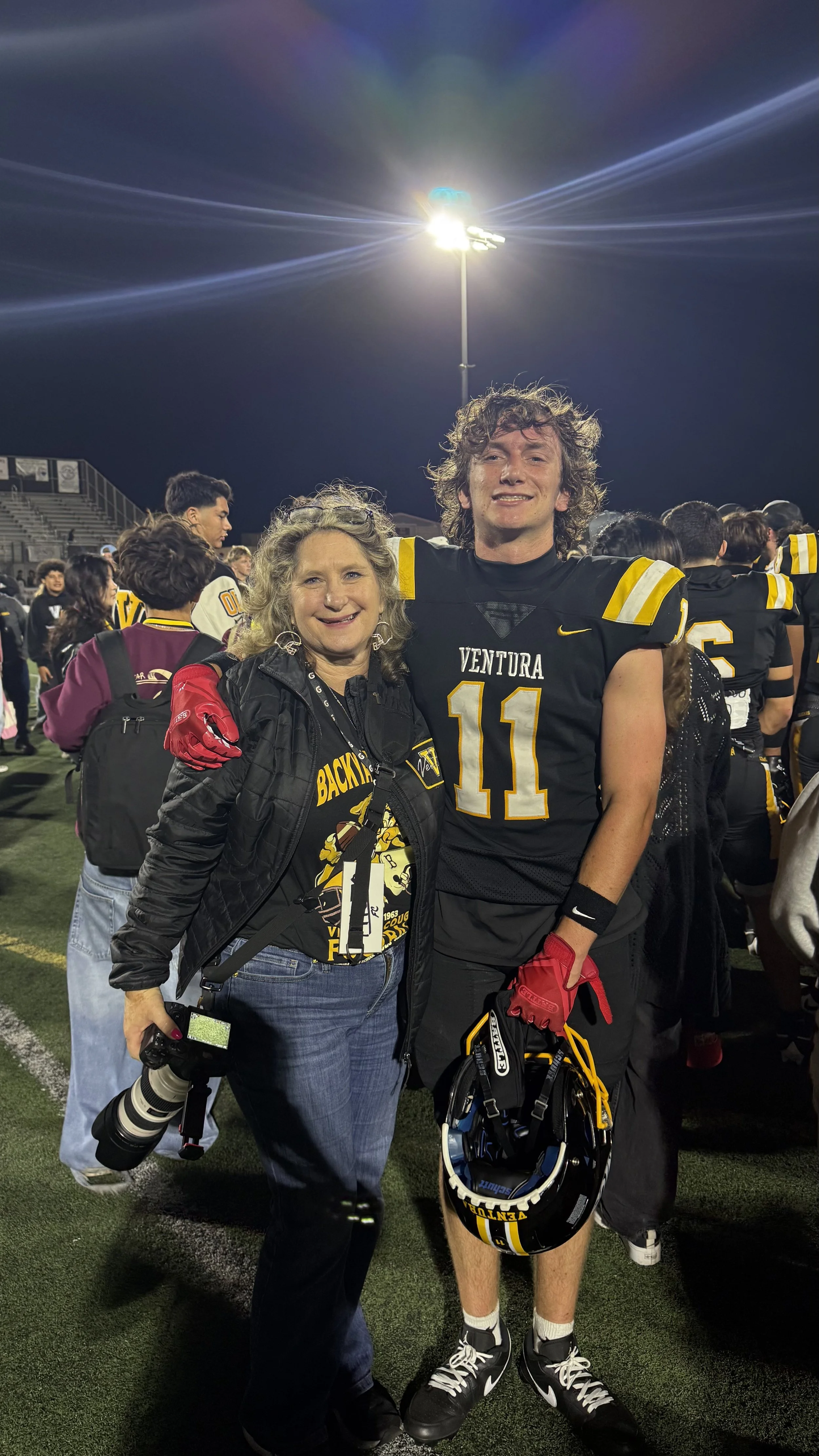 A young football player in black and yellow uniform smiling while holding a helmet, stands beside a woman with gray curly hair in a black jacket, both are on a football field at night with a stadium and bright stadium lights in the background.