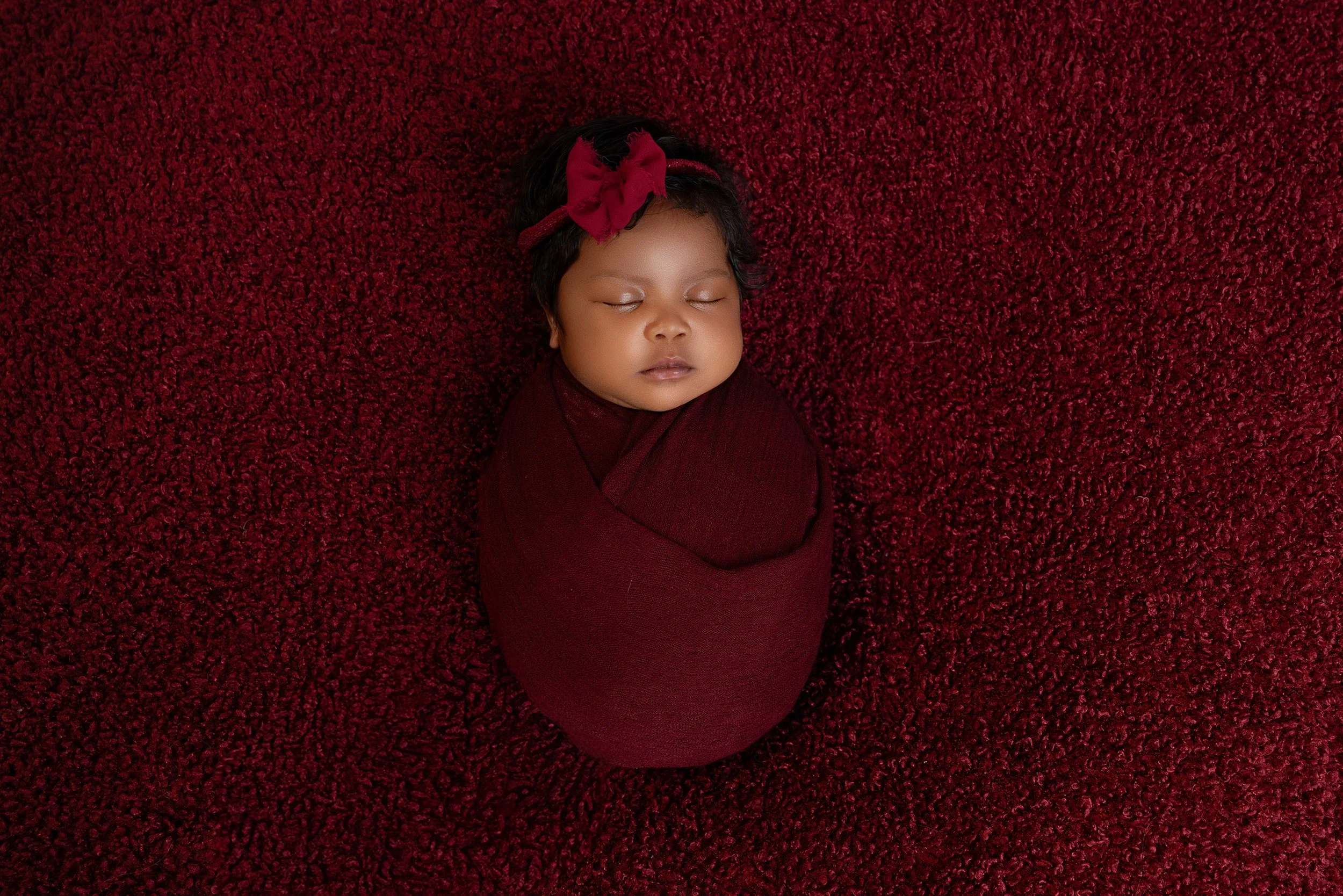 Close-up of a sleeping baby girl with a floral headband, lying on a red textured surface, wearing a maroon outfit.