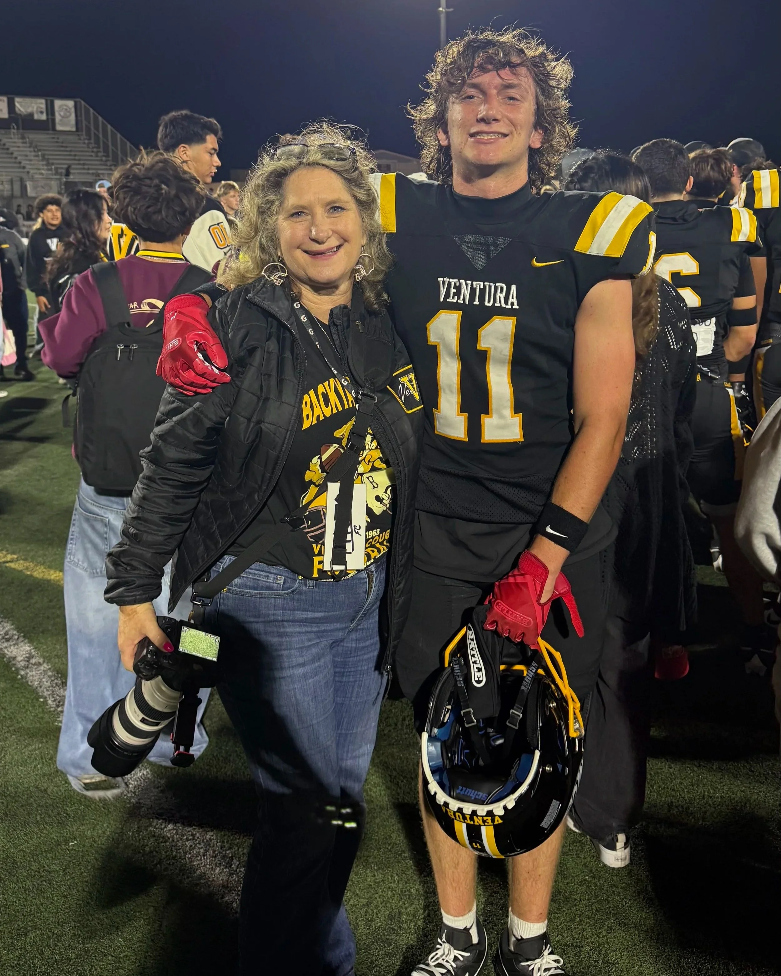 A woman and a football player pose on a football field at night. The woman is holding a camera with a telephoto lens, wearing a black jacket and jeans, and has curly gray hair. The football player is in uniform with a helmet in his hand, wearing a jersey with the number 11 and the name Ventura, and has curly hair.