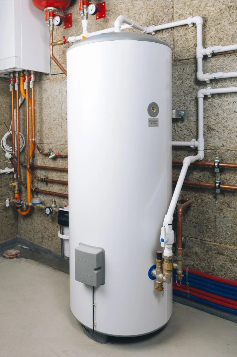 A white, cylindrical water heater in a utility room with pipes attached, surrounded by various valves and gauges.