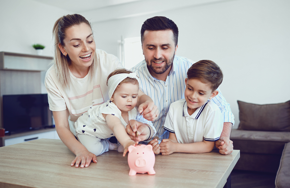 Family with two children playing with a pink piggy bank at a table in a living room.