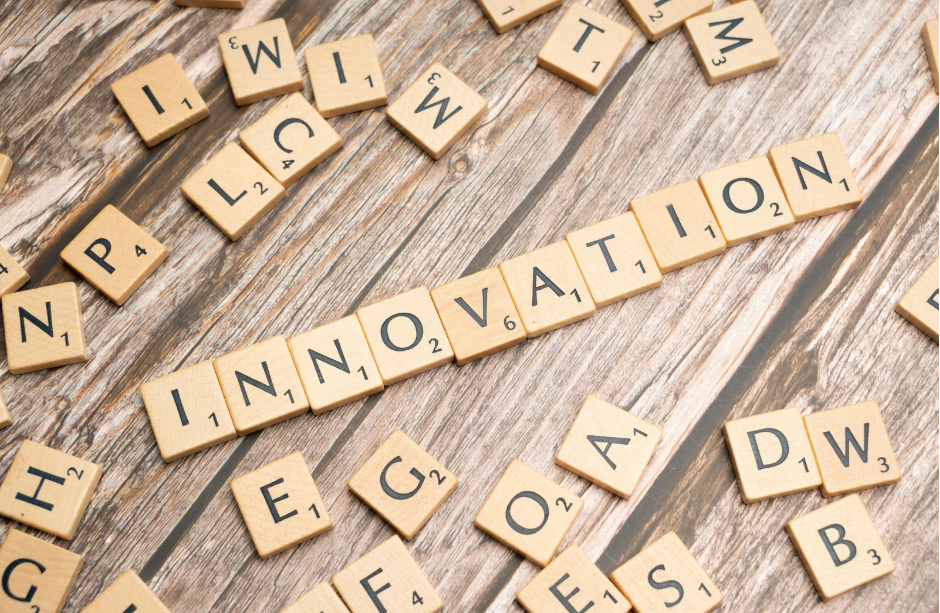 Wooden Scrabble tiles spelling "INNOVATION" on a wooden surface, surrounded by scattered random letters.
