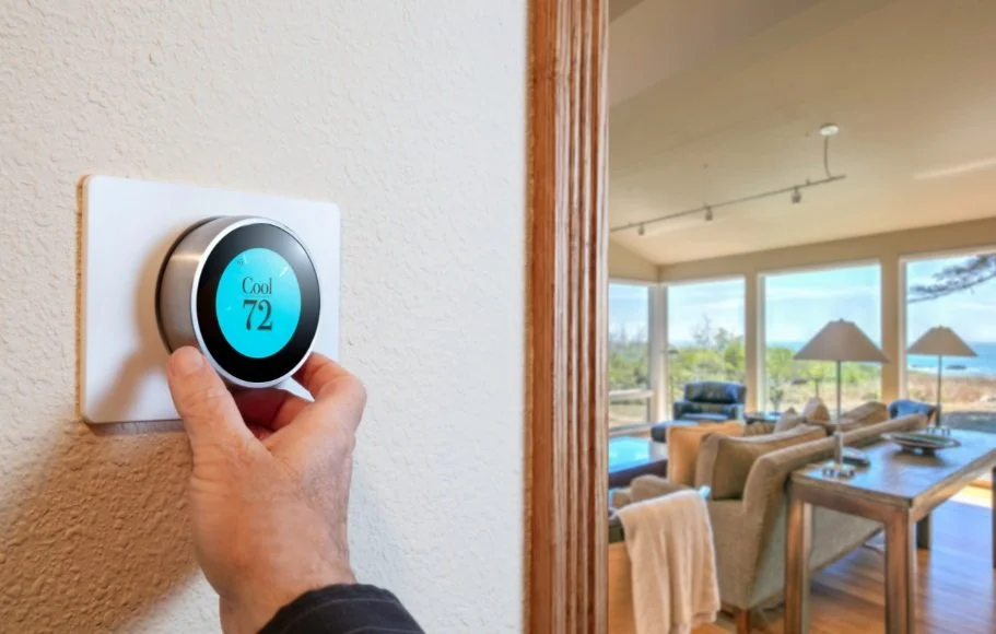Hand adjusting a smart thermostat set to cooling mode at 72 degrees in a contemporary living room with large windows and modern furniture.