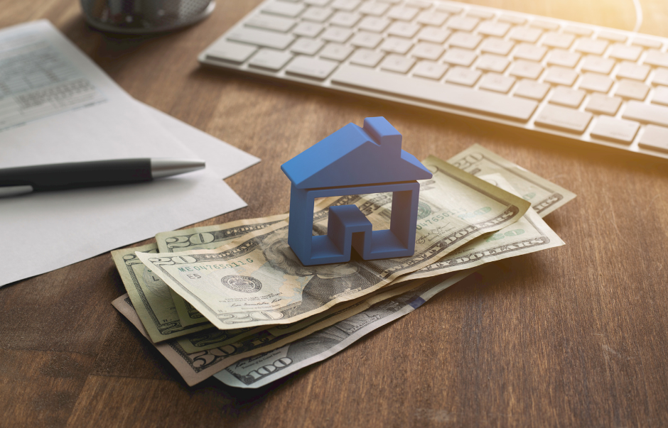 Small blue house model on top of US dollar bills, next to a pen and paper, with a keyboard in the background on a wooden desk.