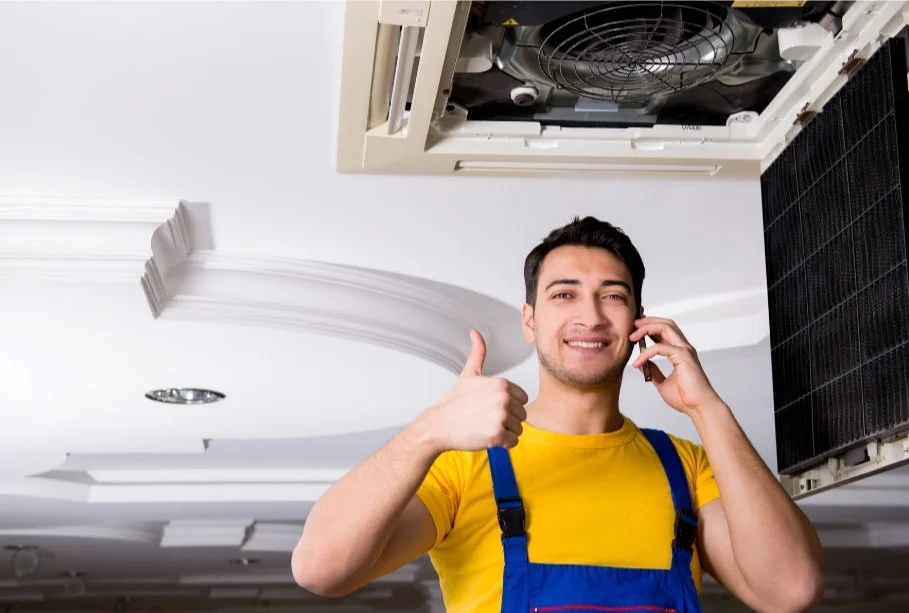 Man in yellow shirt and blue overalls talking on the phone, giving thumbs up, standing near an open ceiling air conditioning unit.
