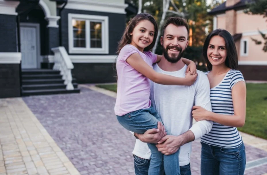 A happy family of three, including a man, woman, and a young girl, standing outside in front of a house. The man is holding the girl, and they are all smiling.