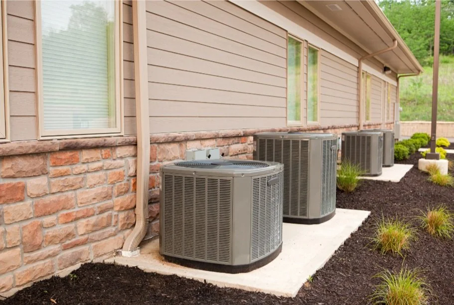 Residential air conditioning units outside a house on concrete pads, surrounded by landscaping, including stone siding and mulch with plants.