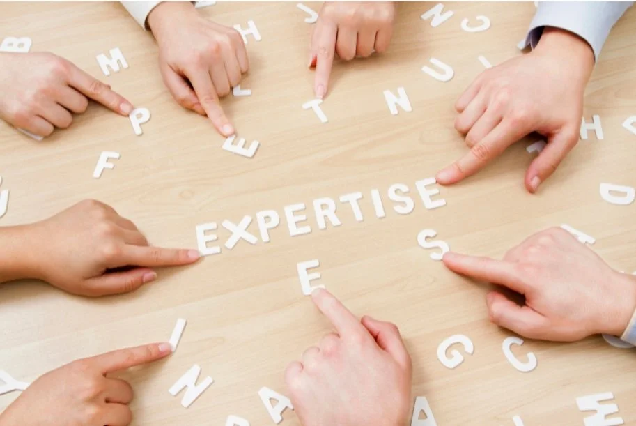 Hands pointing to the word 'EXPERTISE' made of white letters on a wooden surface.