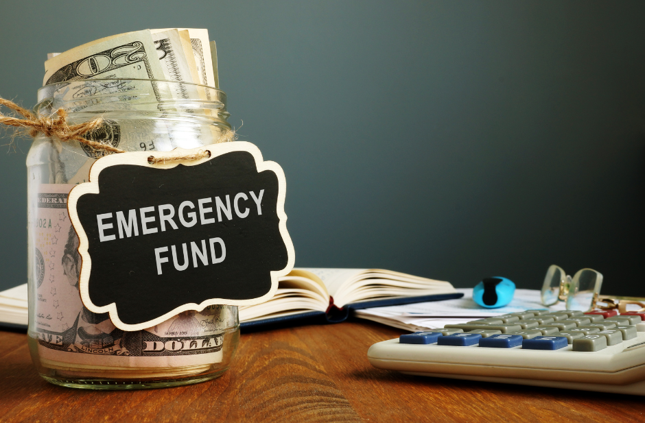Jar labeled 'Emergency Fund' filled with cash, next to a calculator, open notebook, and glasses on a wooden table.
