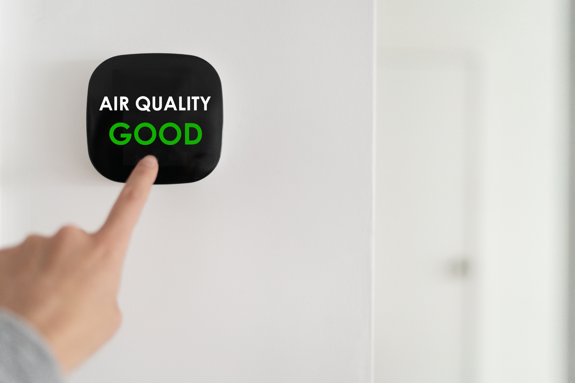 Person's hand pointing at an air quality monitor displaying 'Air Quality Good' on a wall.