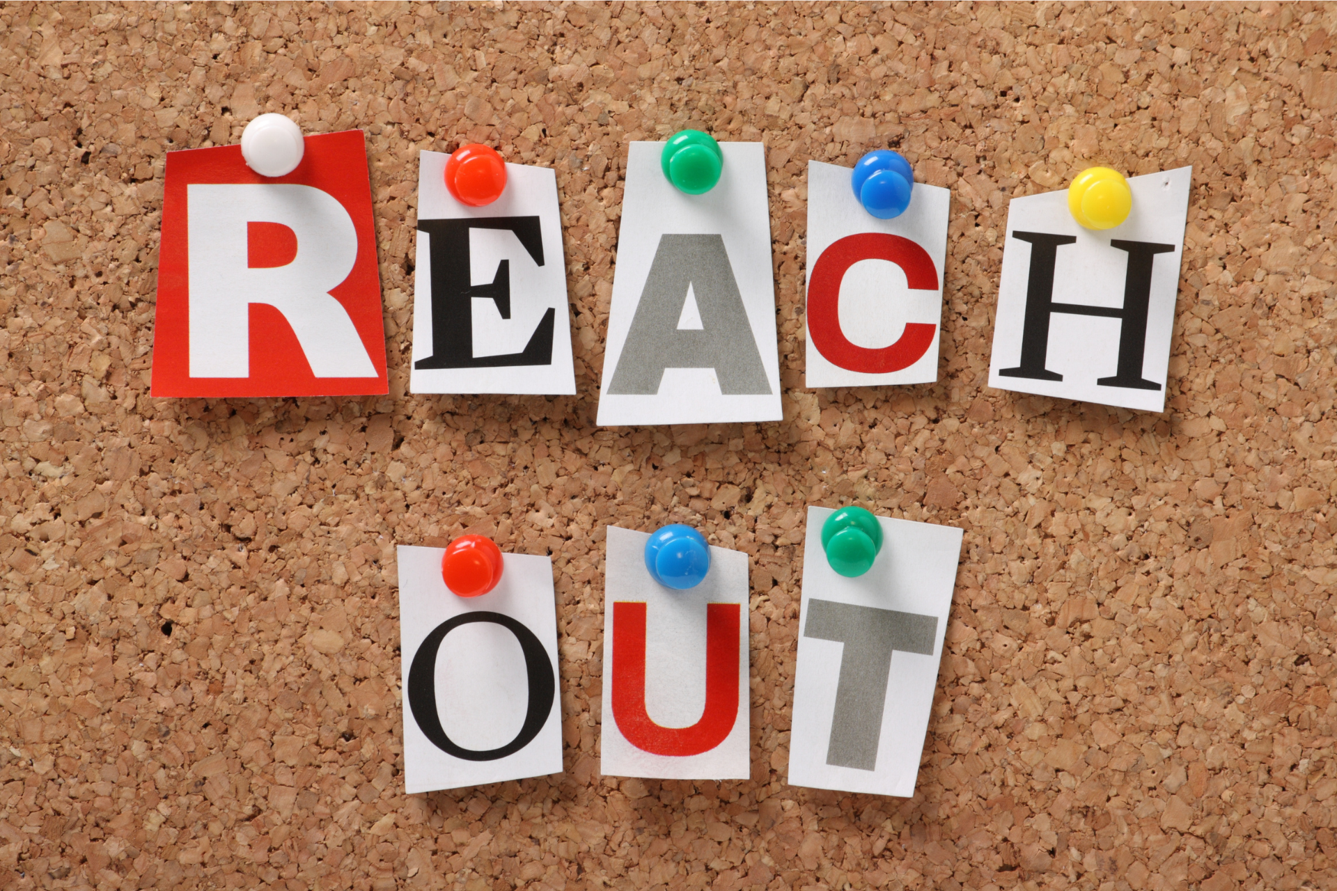 The phrase "REACH OUT" spelled with cut-out letters pinned to a corkboard.