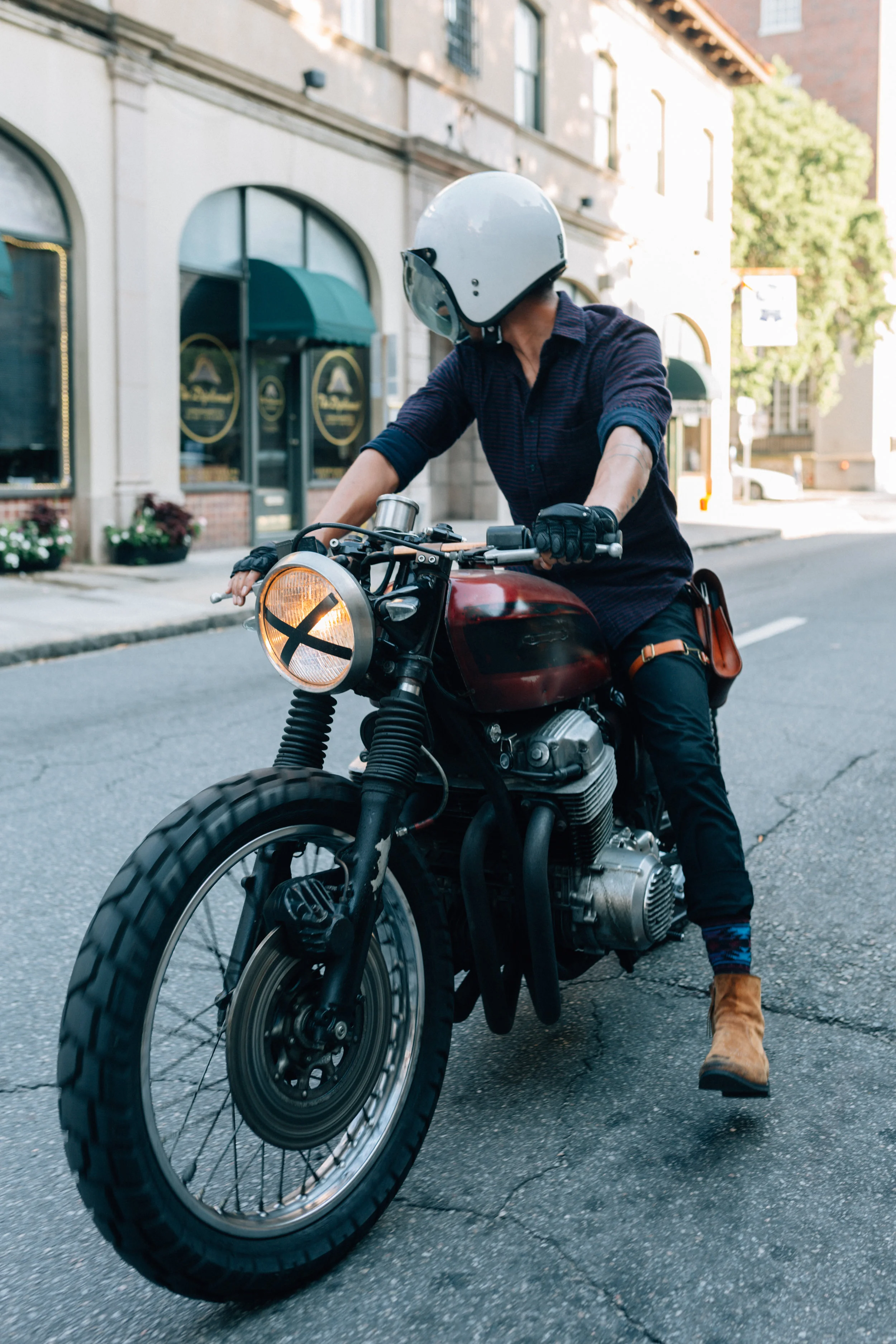 Person riding a motorcycle on city street, wearing a white helmet, dark clothing, and brown boots.