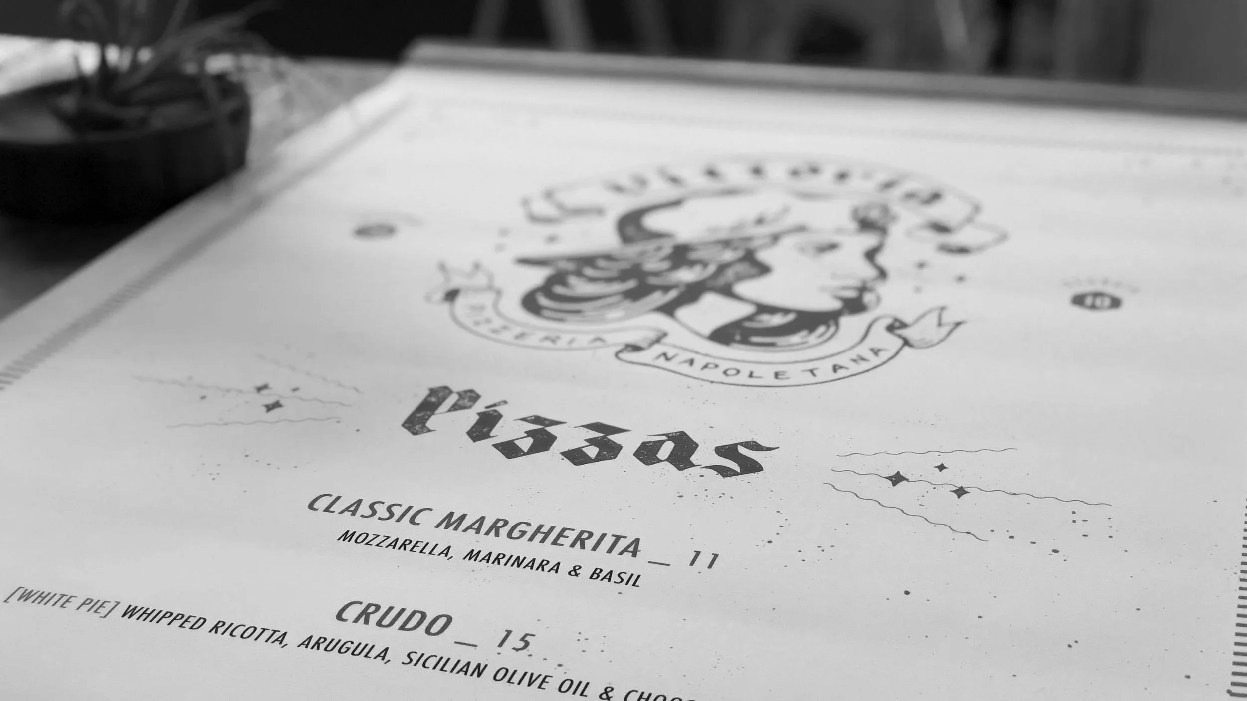 Black and white photo of a pizza restaurant menu with names and prices, featuring a decorative logo at the top.