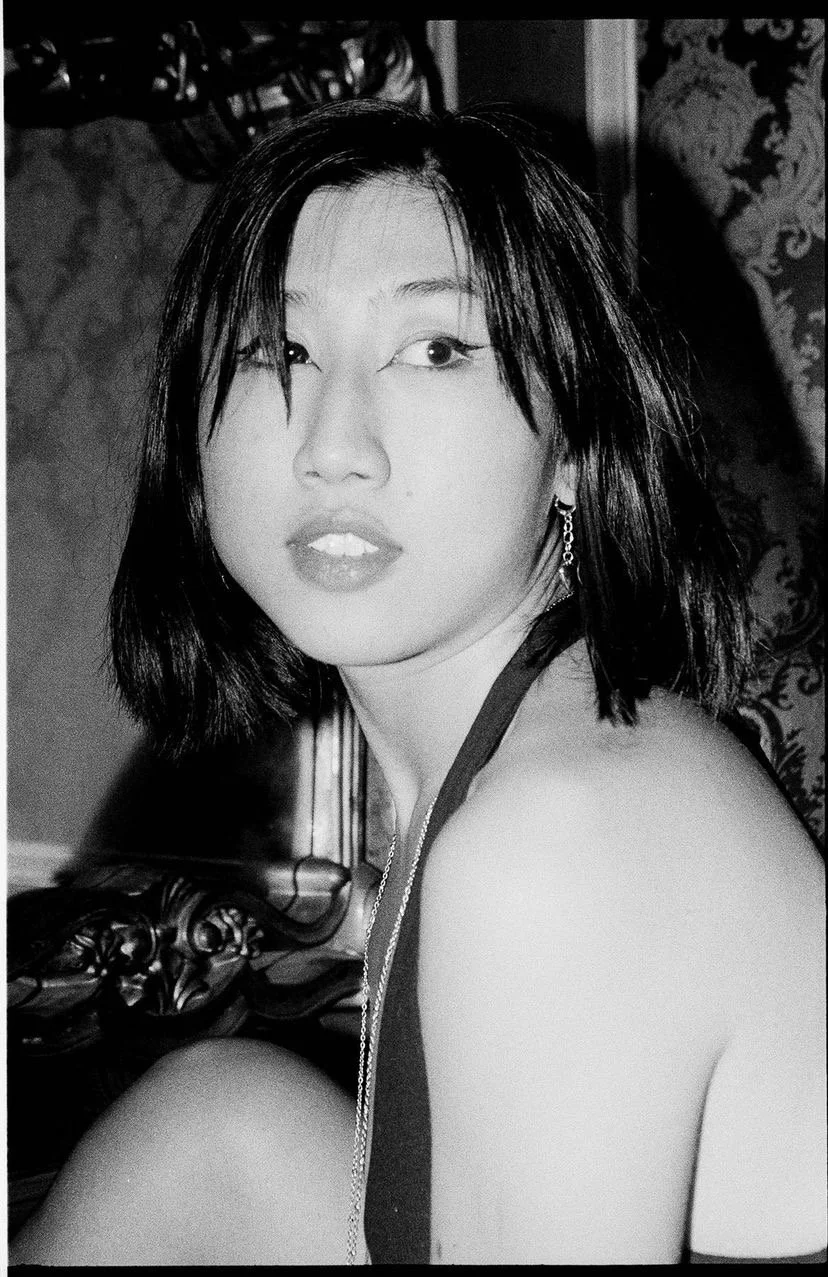 Black and white photograph of a woman with shoulder-length dark hair, wearing earrings and a sleeveless top, looking over her shoulder with a slightly parted lips expression.