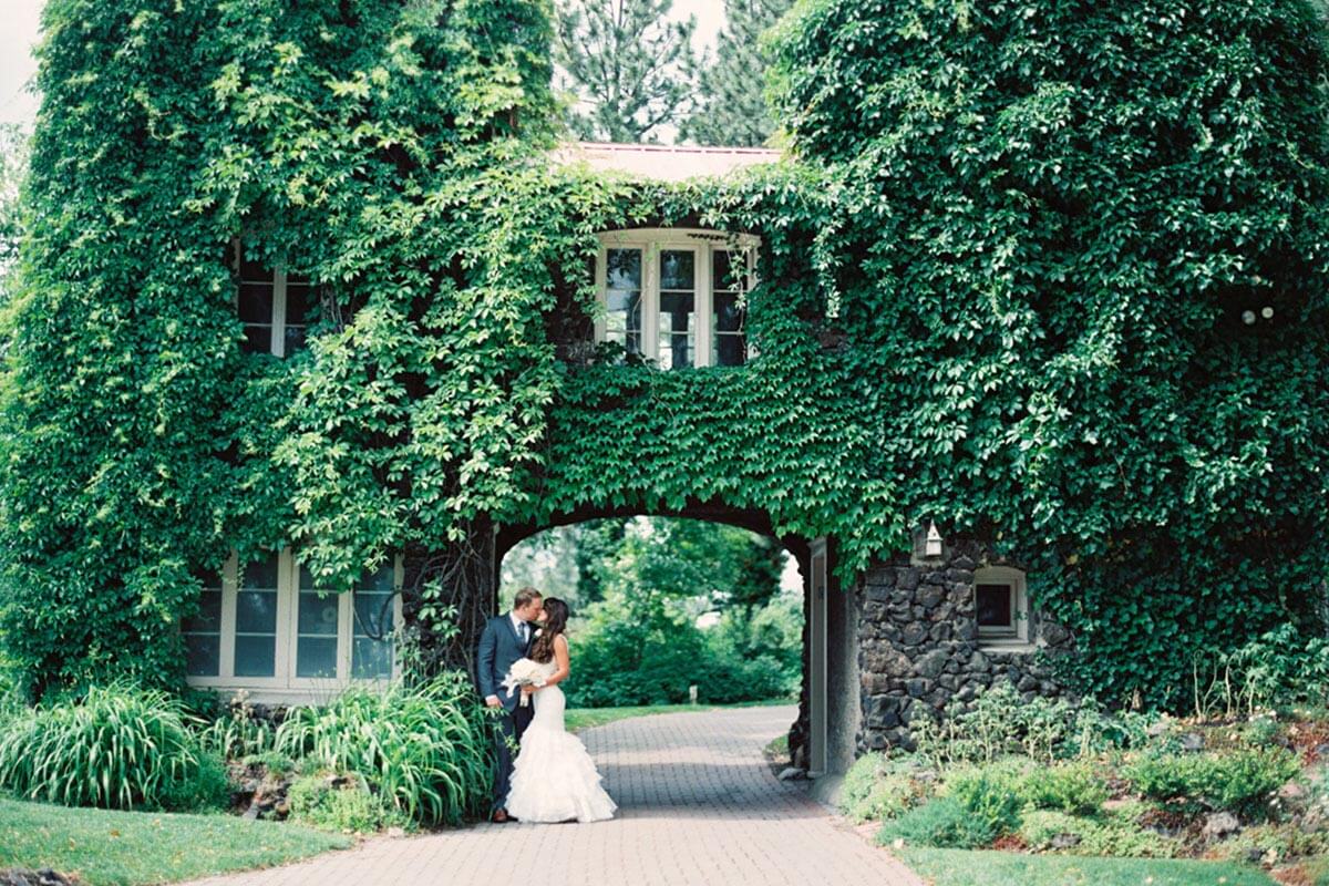 A photo of a bride and groom kissing in front of the gatehouse with overgrown greenery