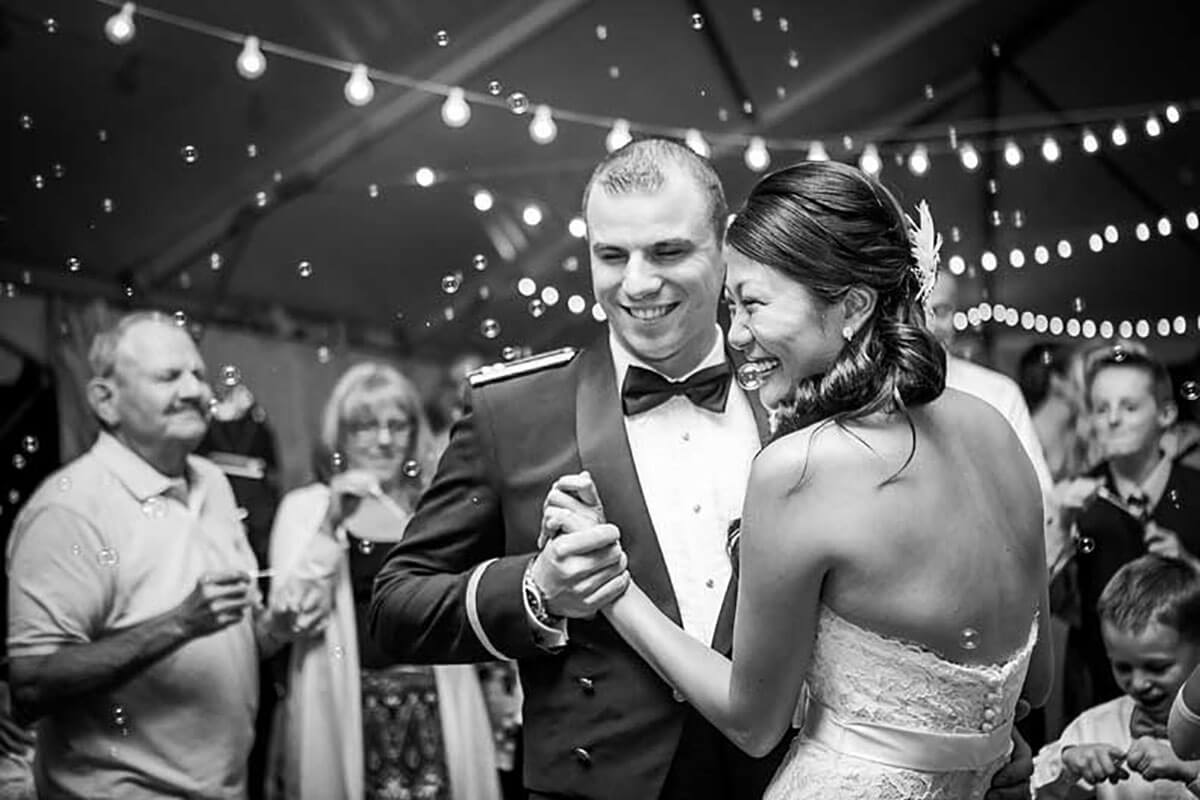 A black and white photo of a bride and groom smiling as they dance in the wedding tent with friends and friends behind them 