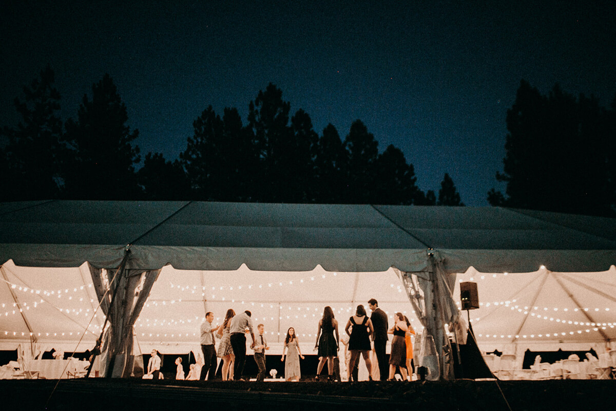 A night time shot of the wedding tent exterior with wedding party inside. Trees frame the photo against a night sky. 
