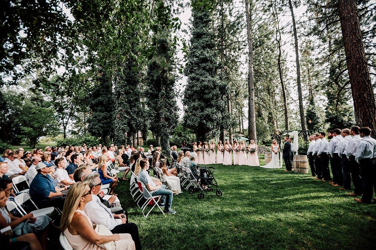 A photo of a wedding ceremony in the pine trees with crowd sitting to the left and the wedding party to the right