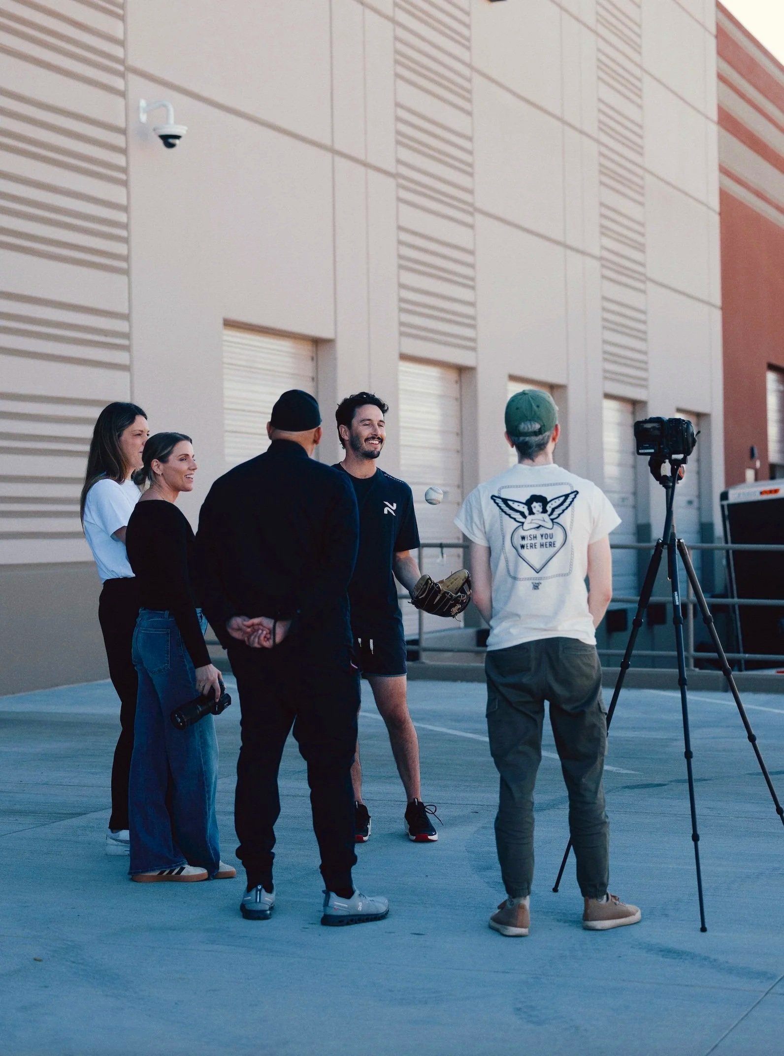 A group of five people standing outdoors, engaged in conversation with a camera and microphone set up nearby. The scene takes place in front of a beige building with horizontal and vertical siding patterns.