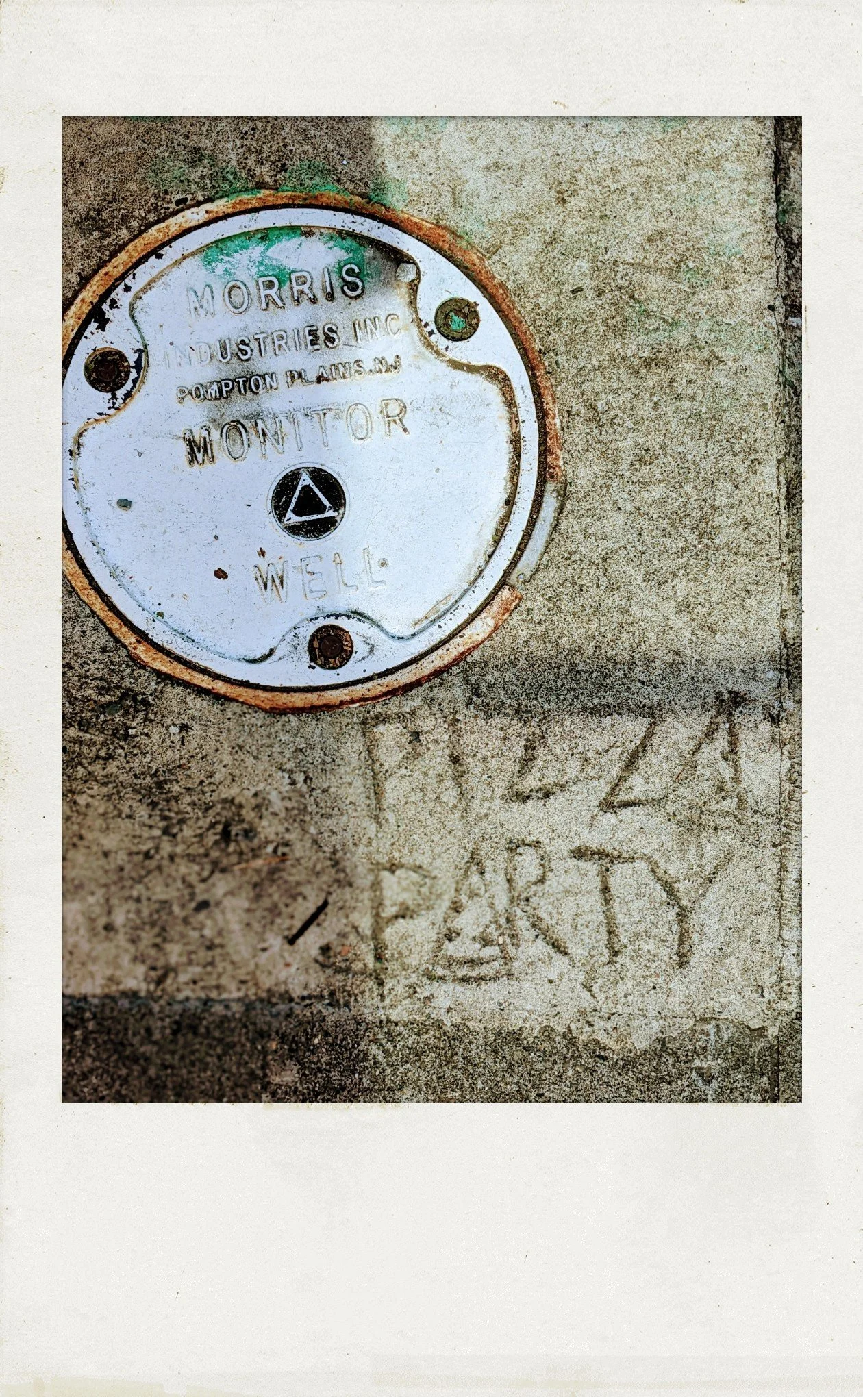 photograph in polaroid frame, saturated color, high-contrast. looking down at the sidewalk: upper left corner, a round metal plate, similar to a manhole cover. lower right, someone has etched "PIZZA PARTY" in the concrete.