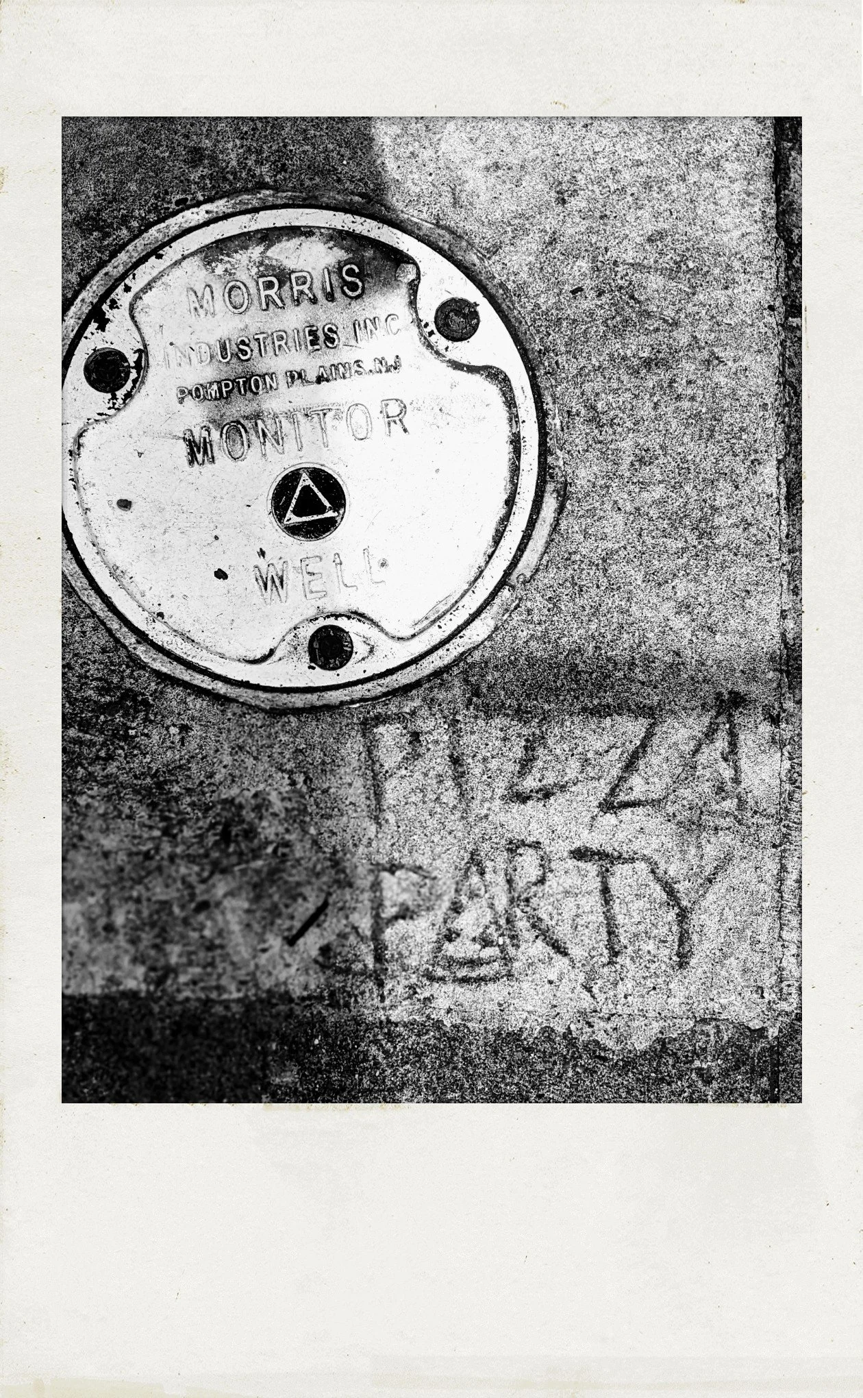 photograph in polaroid frame, black & white. looking down at the sidewalk: upper left corner, a round metal plate, similar to a manhole cover. lower right, someone has etched "PIZZA PARTY" in the concrete.