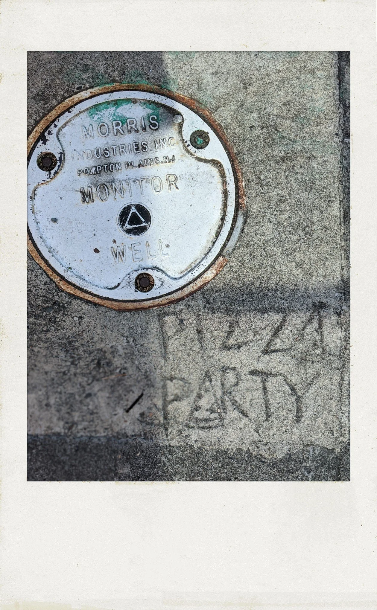 photograph in polaroid frame, natural light. looking down at the sidewalk: upper left corner, a round metal plate, similar to a manhole cover. lower right, someone has etched "PIZZA PARTY" in the concrete.