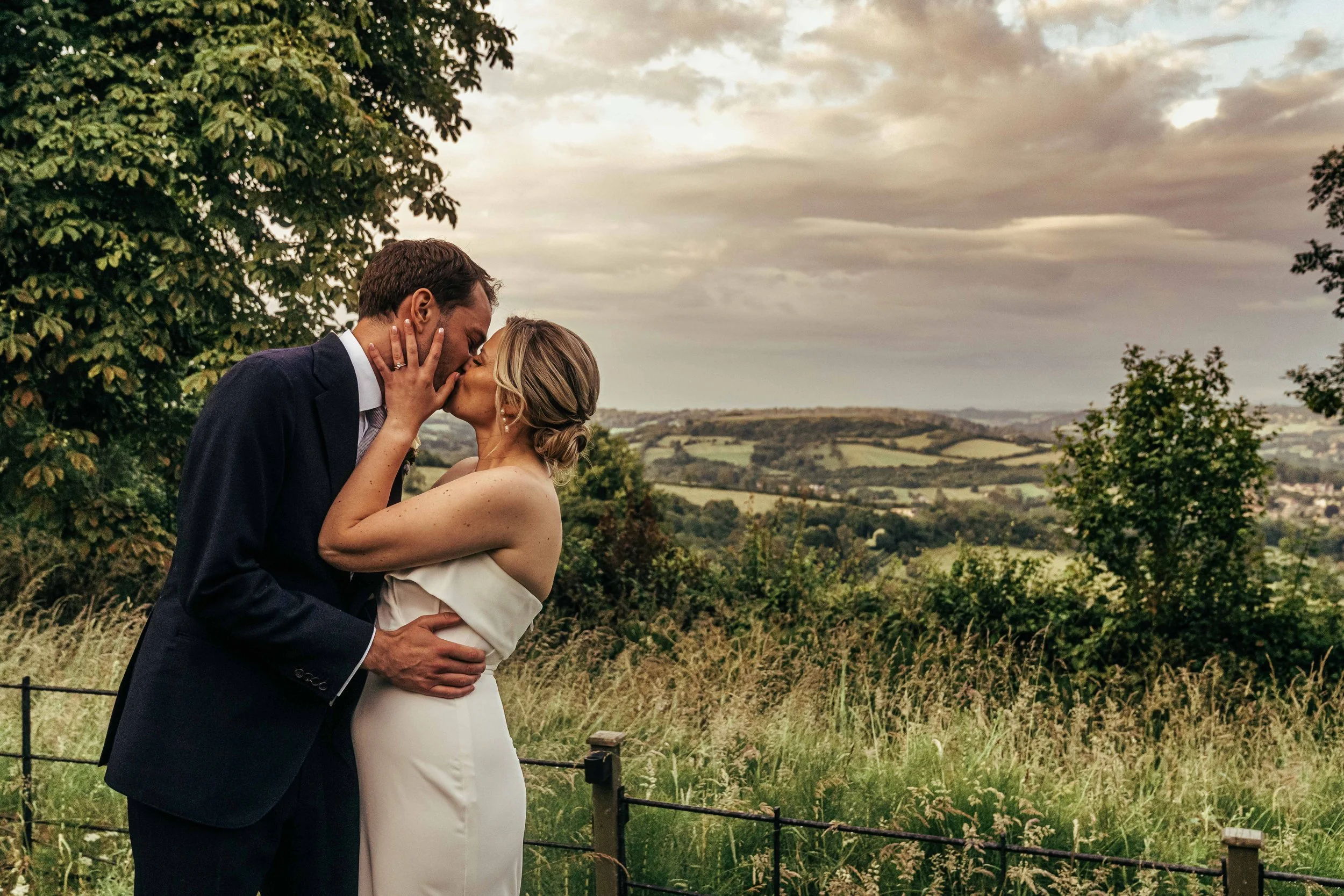 A couple sharing a kiss outdoors in a scenic, countryside setting during the daytime. The man is wearing a dark suit, and the woman is in a white off-the-shoulder dress. They are embracing each other with a beautiful landscape of rolling hills and a cloudy sky in the background.