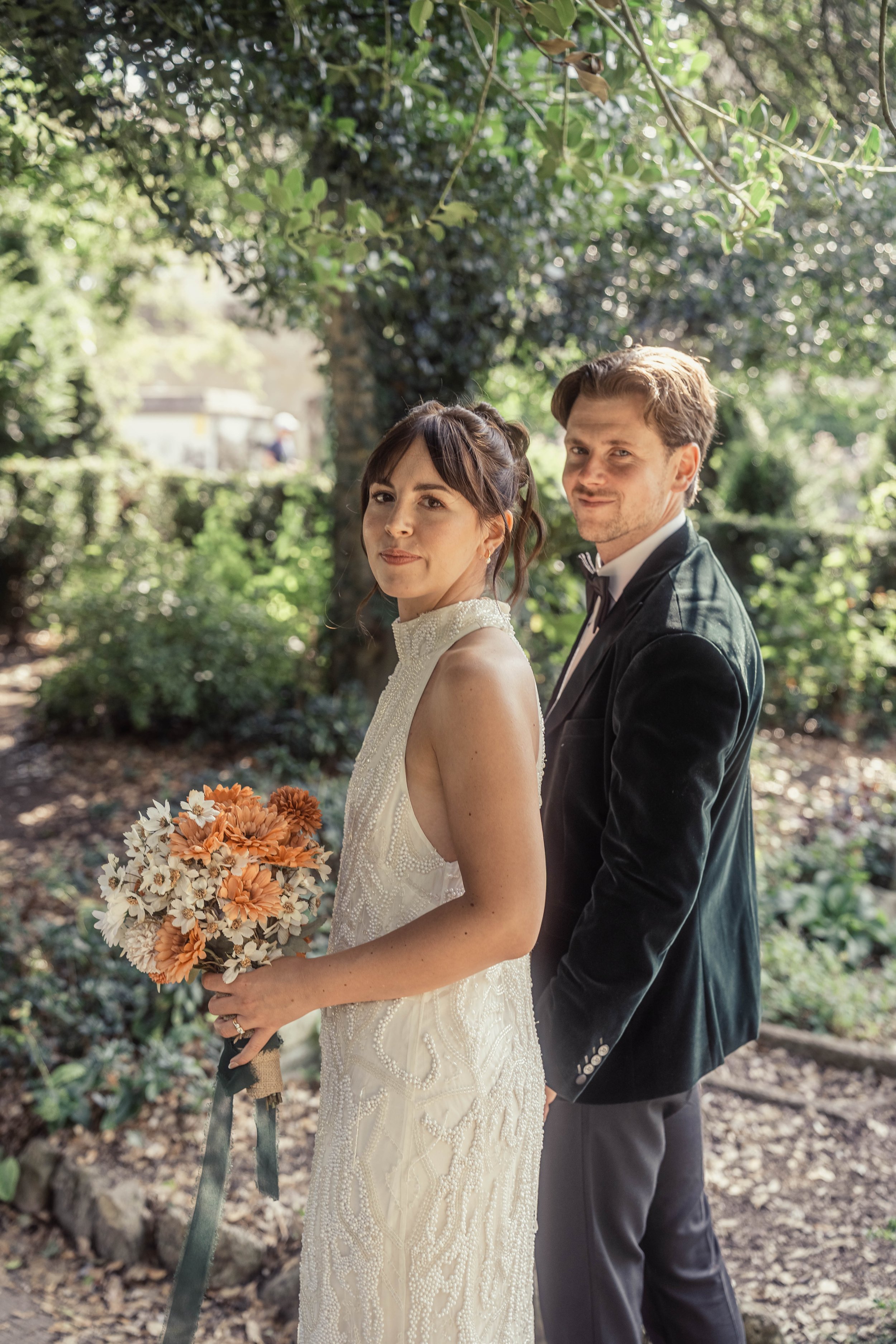 A bride in a white beaded gown holding a bouquet of light orange and white flowers, standing next to a groom in a black tuxedo with a bow tie, in a lush, green outdoor setting.