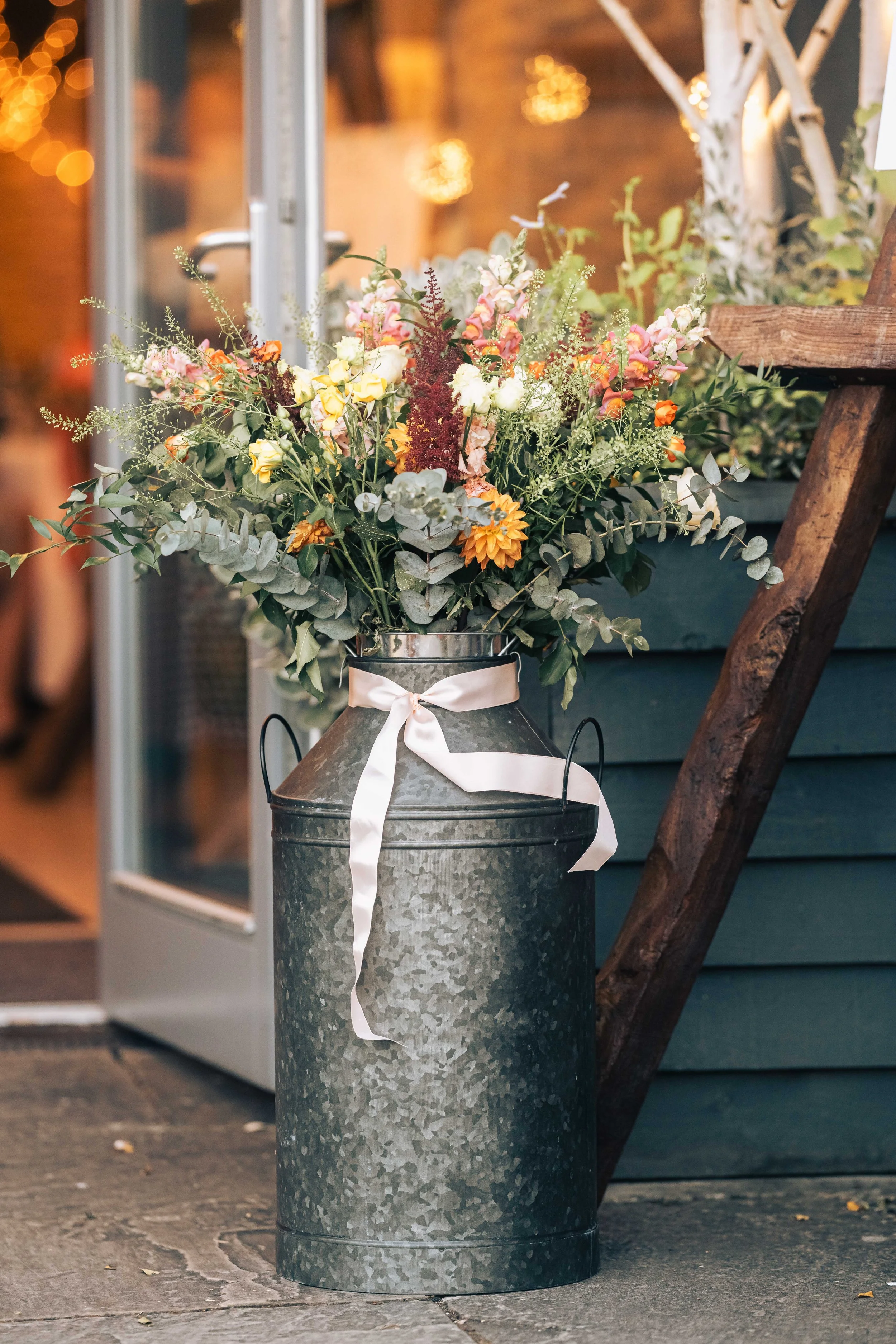 A large metal trash can with a white ribbon tied around it, holding a vibrant bouquet of mixed flowers including white, yellow, orange, and pink blossoms, placed outside a building with a glass door and wooden stairs.