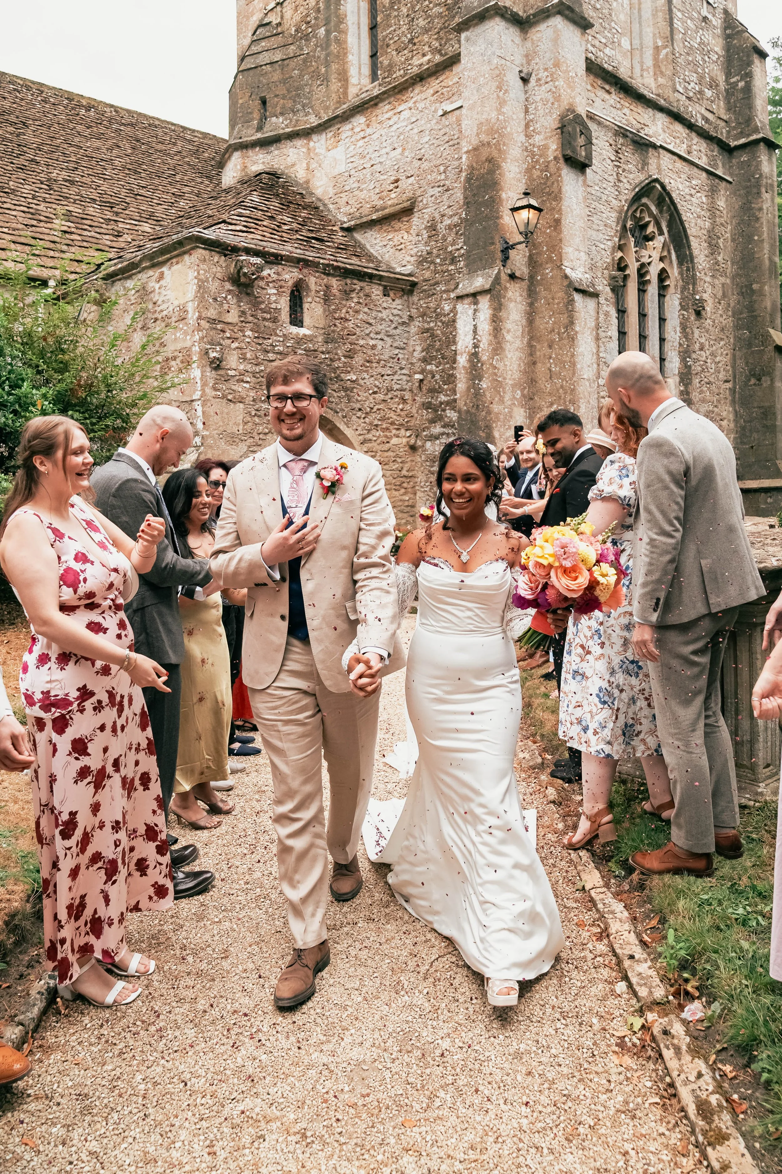 A wedding celebration outside a historic stone church with a diverse group of guests, including a bride in a white gown and a groom in a beige suit, holding hands and smiling, surrounded by friends and family dressed in colorful formal attire.