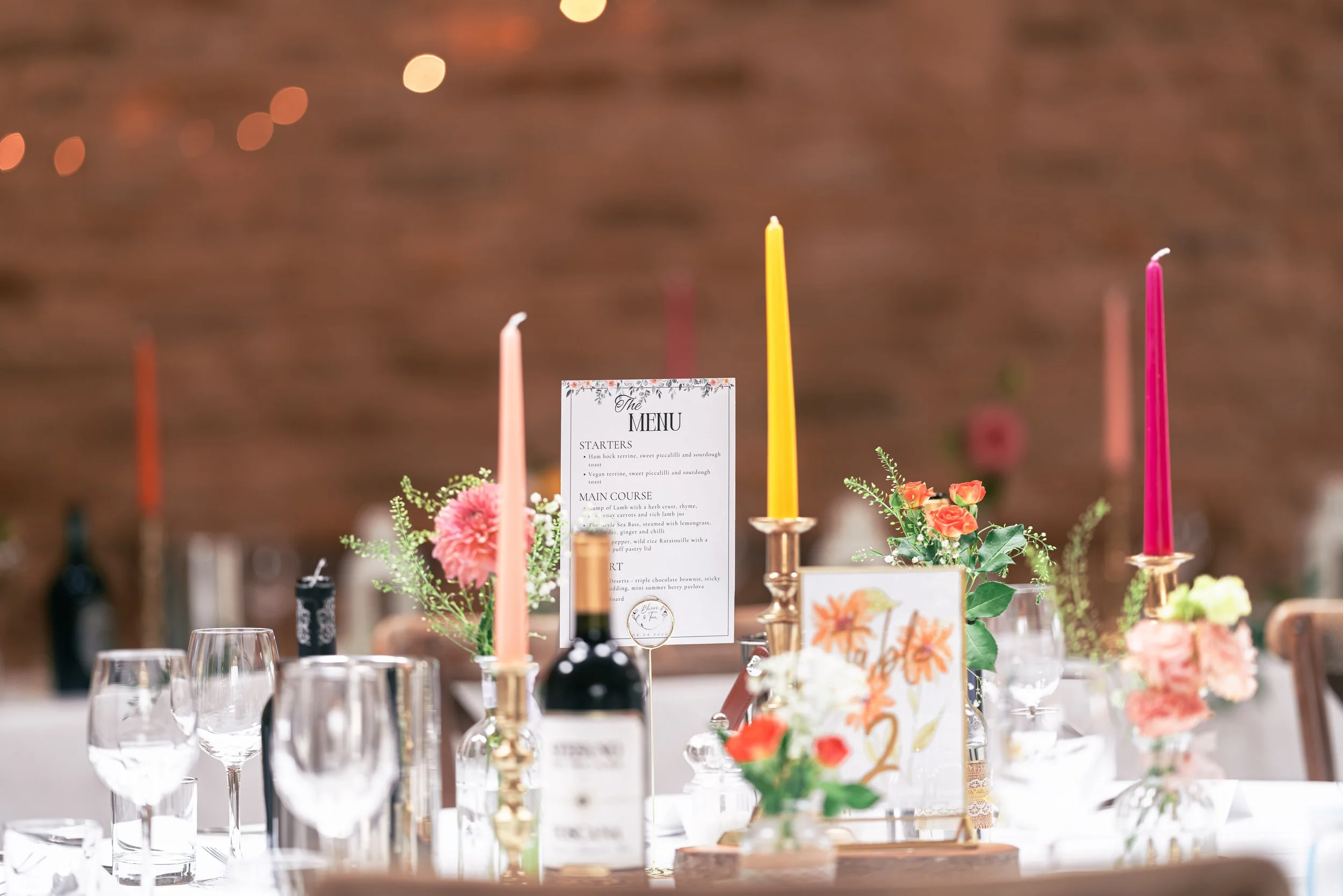 A wedding or event table setup with various colorful candles, flowers, a menu card, wine bottles, and glassware, with a blurred brown background.