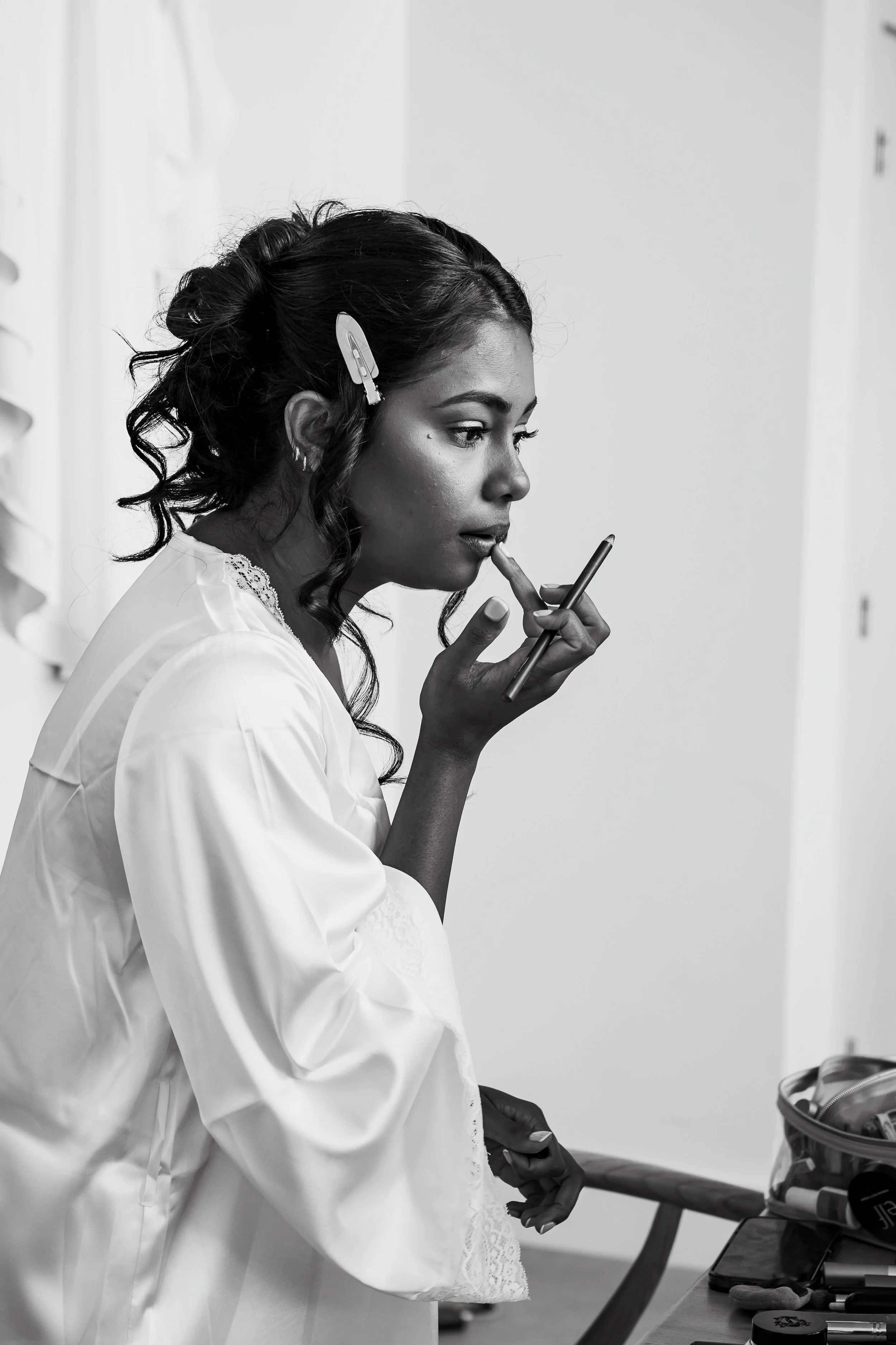 A woman in a white dress, possibly a bride, is examining her finger while holding a smartphone. She is in a room with a table that has makeup and beauty products, and a mirror in the background.