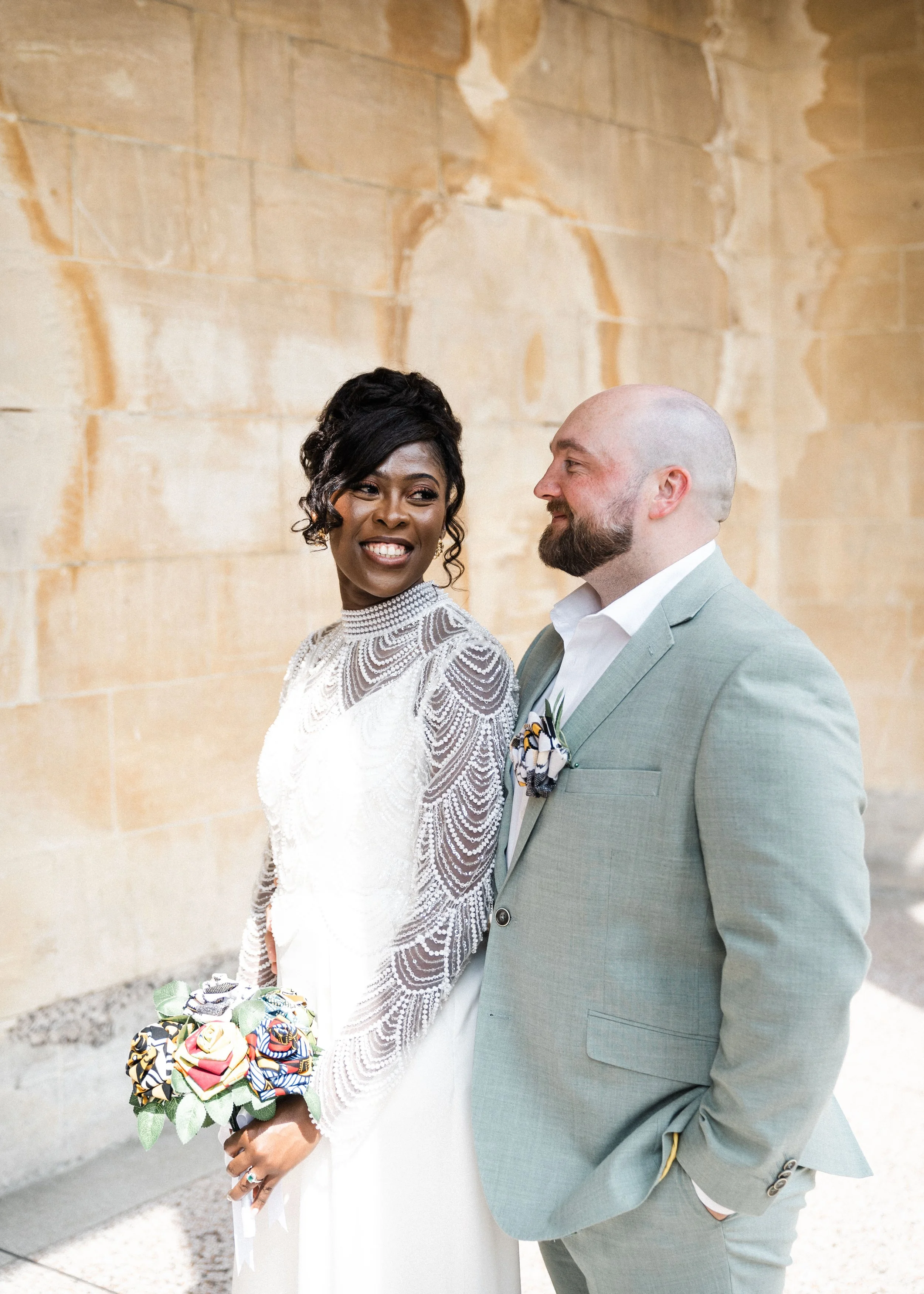 A smiling woman in a white lace wedding dress holding a bouquet of colorful fabric flowers, standing next to a man in a light gray suit, outdoors against a beige stone wall.