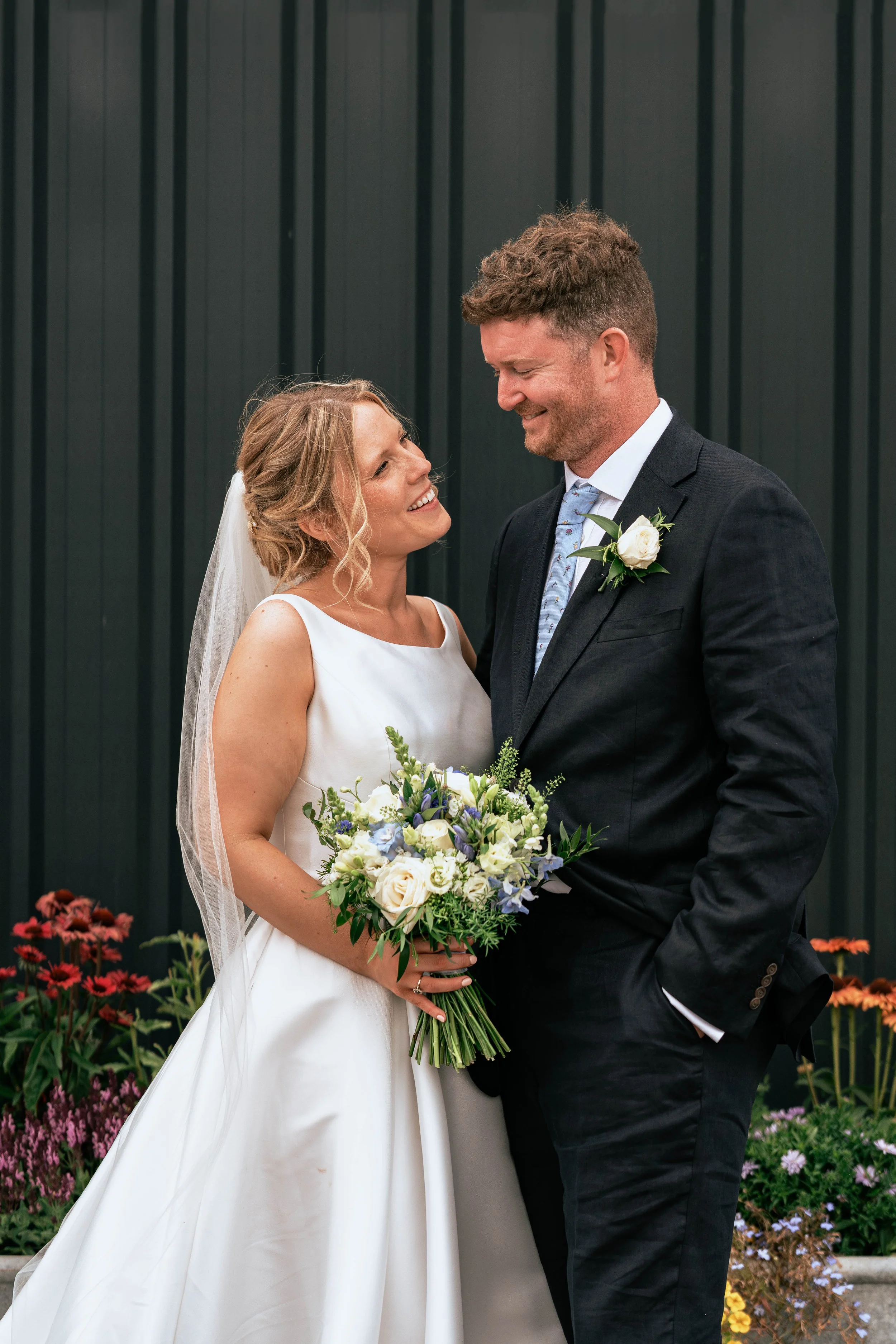A bride and groom smiling and looking at each other during their wedding, with the bride holding a bouquet of flowers and wearing a white dress, and the groom wearing a black suit with a boutonniere, standing in front of a dark green wall with colorf