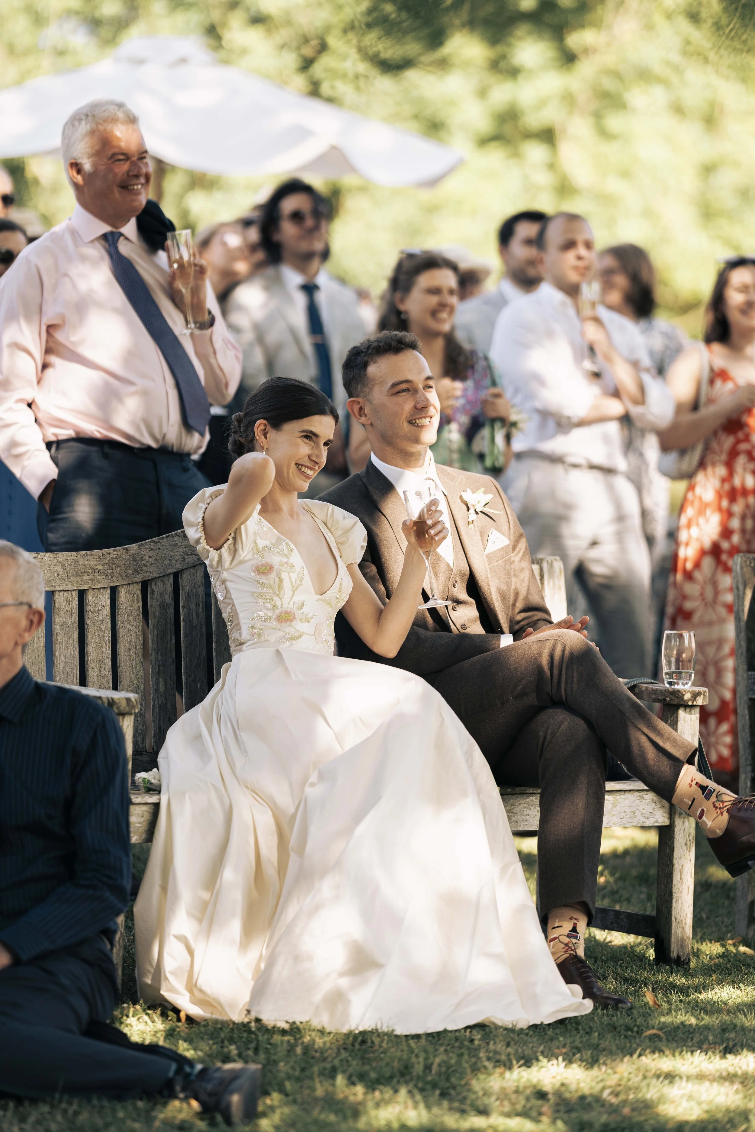 People gathered outdoors for a wedding reception, with a bride and groom sitting on a wooden bench, smiling and holding glasses, surrounded by guests.