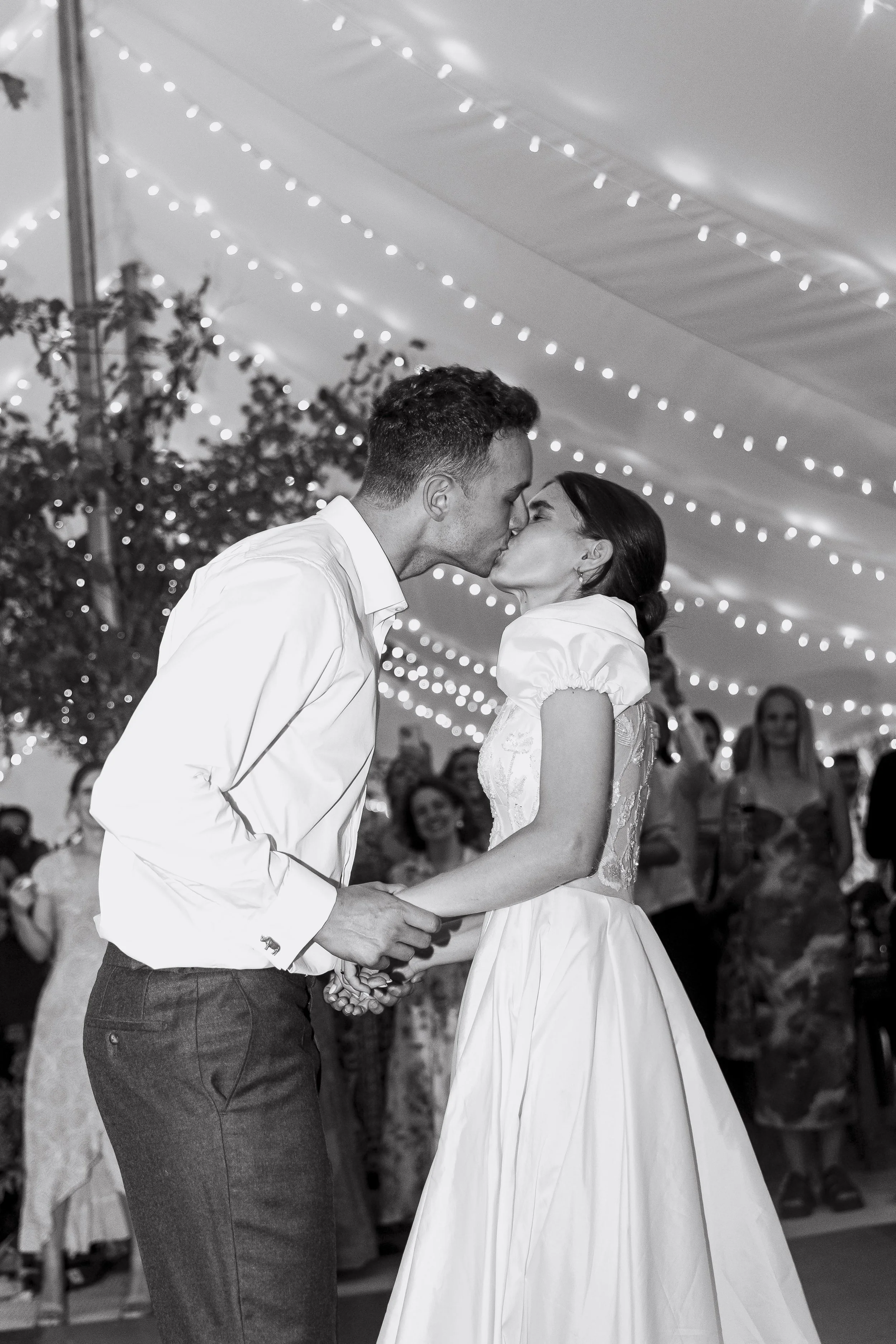 A couple is kissing during their wedding ceremony, holding hands, under string lights with guests watching in the background.