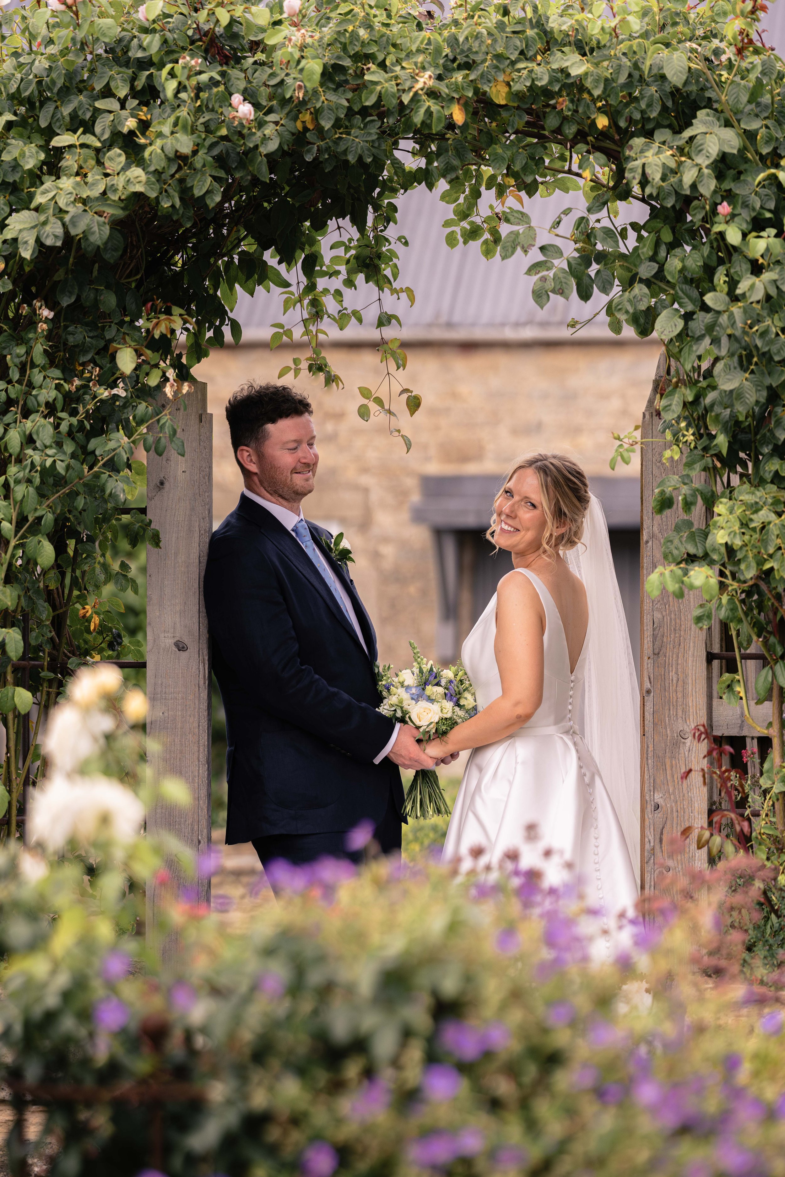 A bride and groom standing beneath a wooden archway decorated with lush green leaves at an outdoor wedding ceremony, smiling and holding hands.