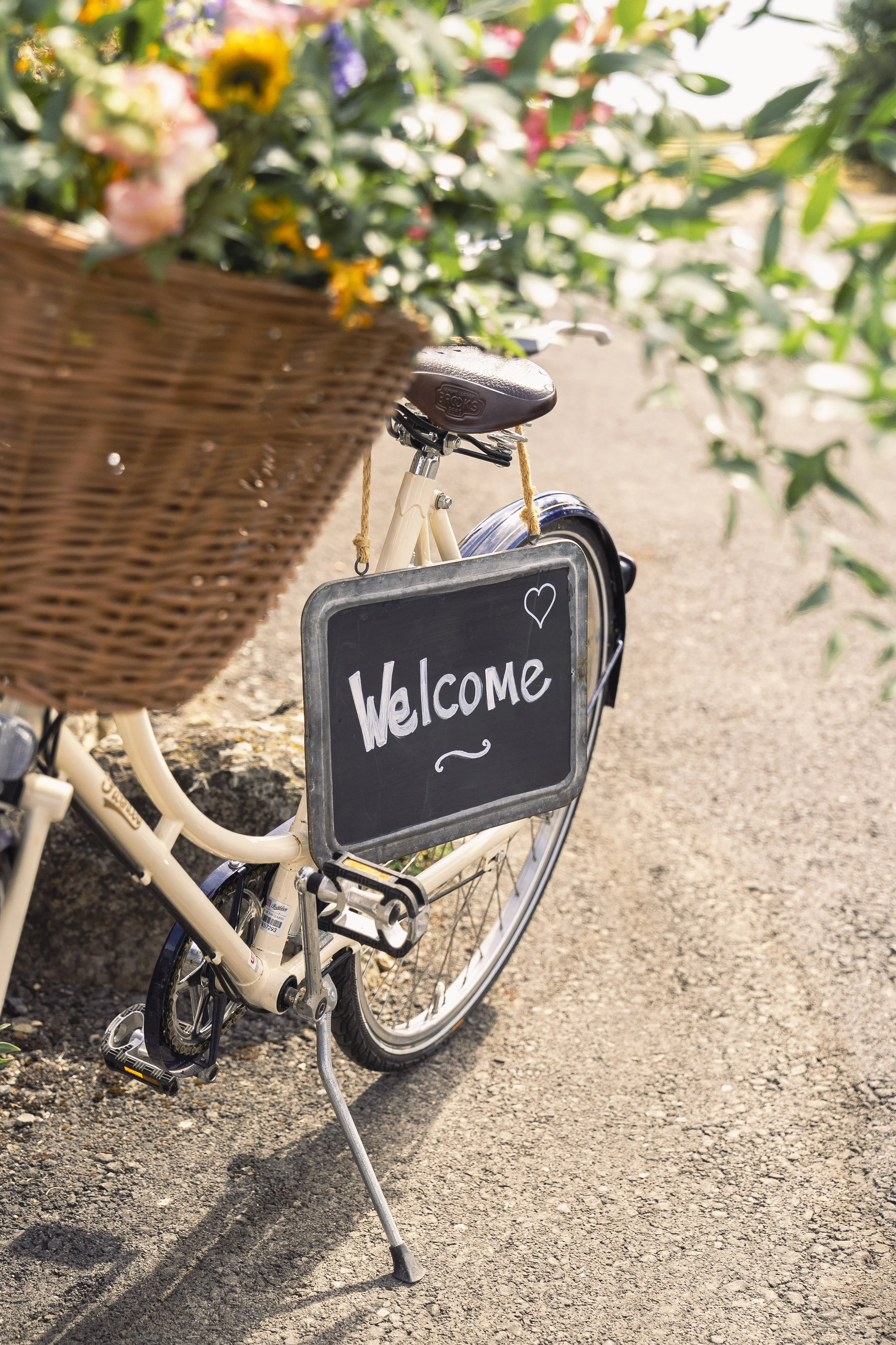 A cream-colored bicycle with a wicker basket filled with colorful flowers in the front. A chalkboard sign attached to the bike reads 'Welcome' with a heart symbol. The bike is on a paved path with greenery and trees in the background.