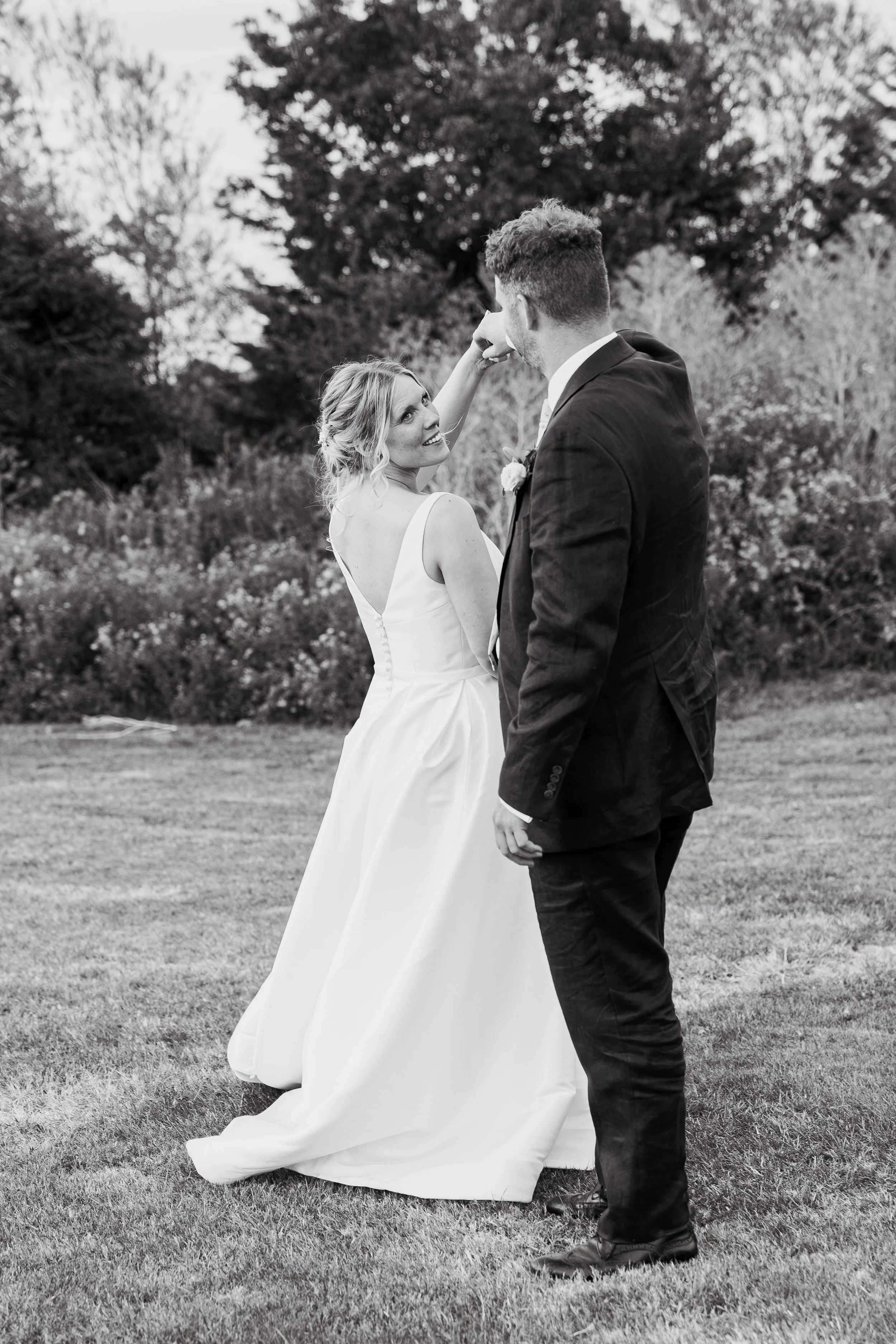 A bride and groom standing on a grassy field during an outdoor wedding, holding hands and looking at each other, with trees in the background.