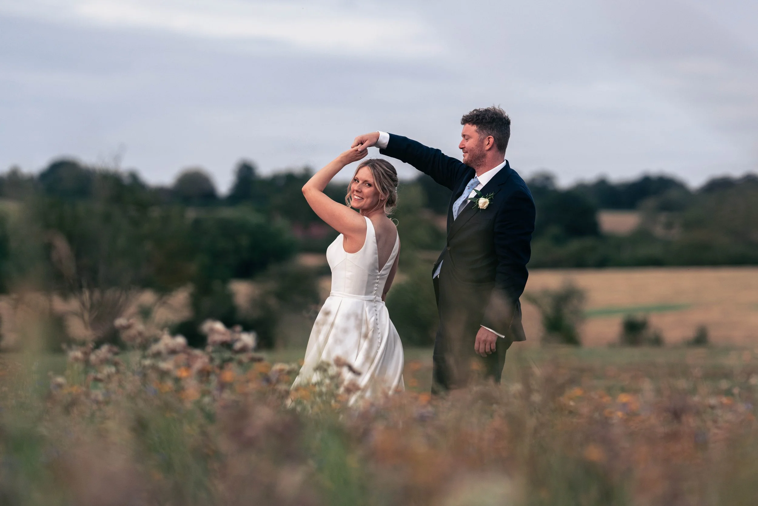 A newlywed couple dancing outdoors in a field, with the groom twirling the bride. The bride is smiling and wearing a white wedding dress, while the groom is dressed in a dark suit with a boutonniere.