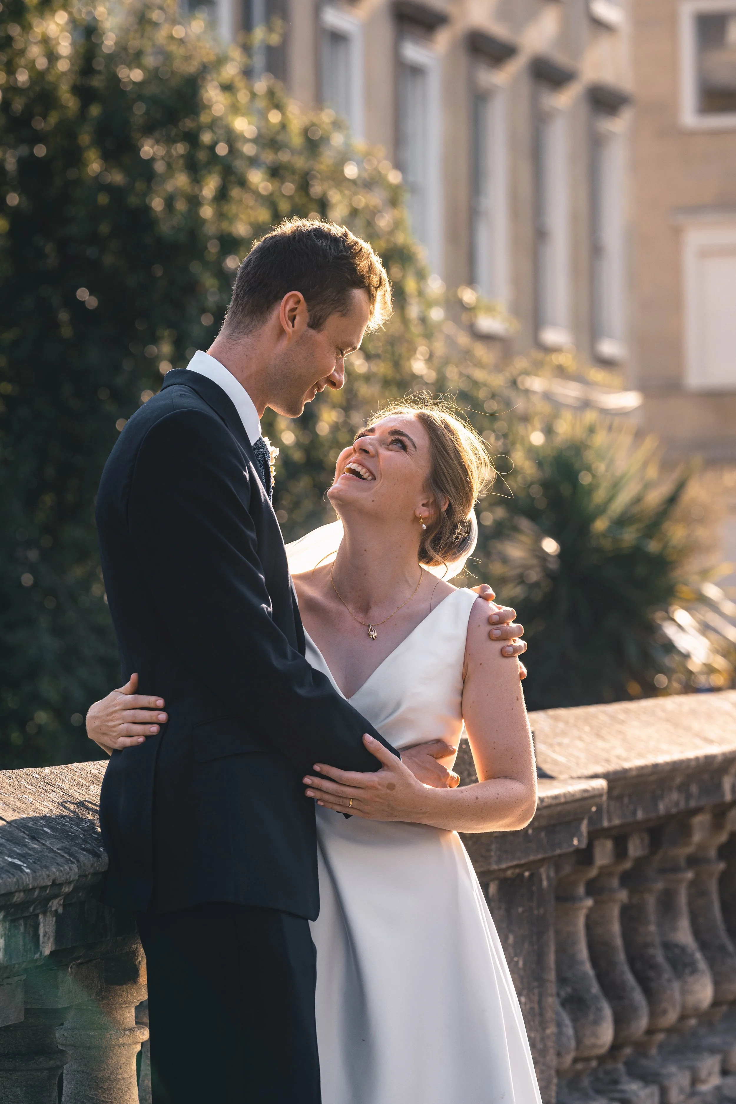 A couple dressed in wedding attire smiling and gazing at each other on a bridge during sunset.