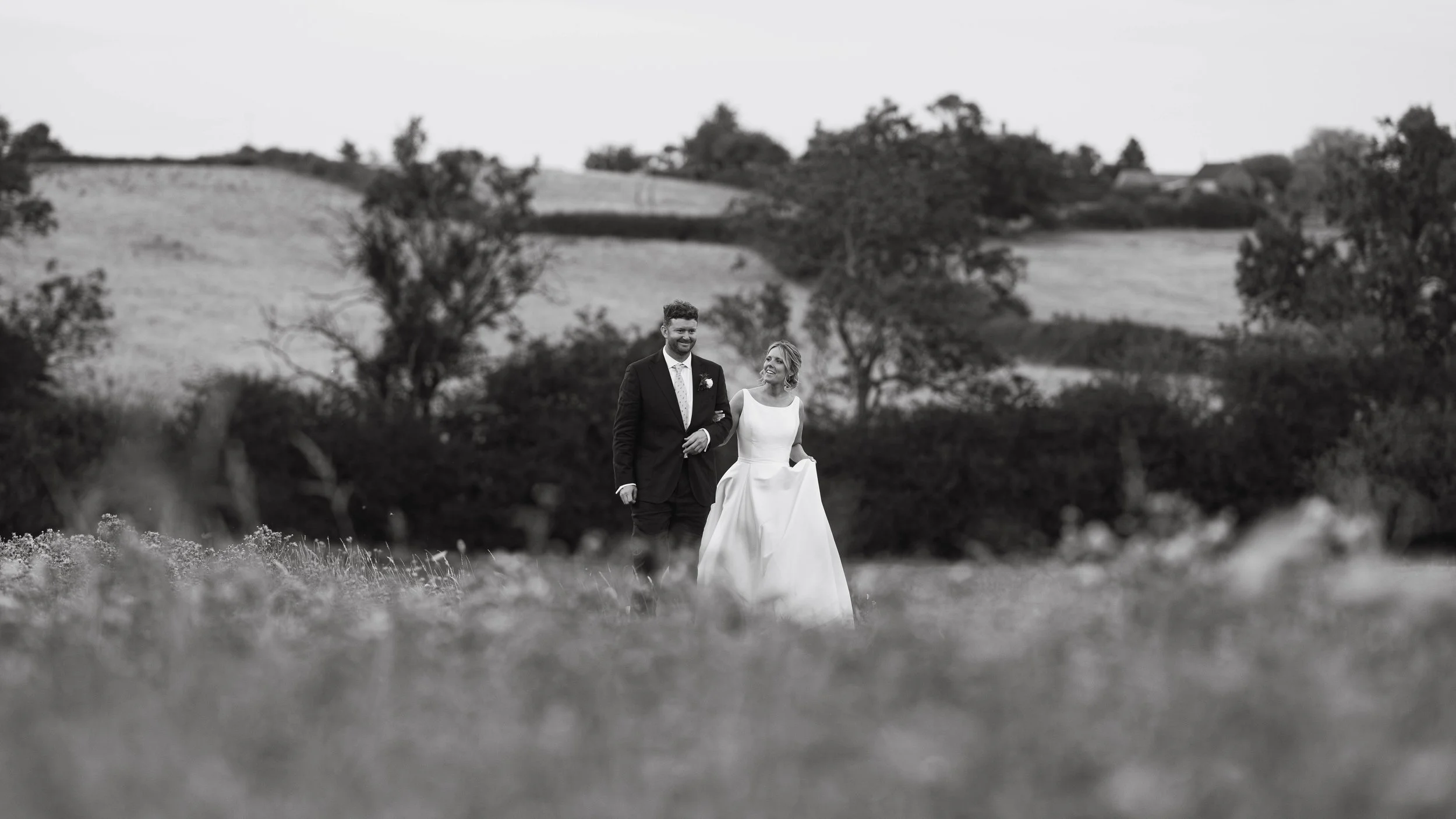 A black-and-white photograph of a bride and groom walking arm in arm through a field with trees and rolling hills in the background.